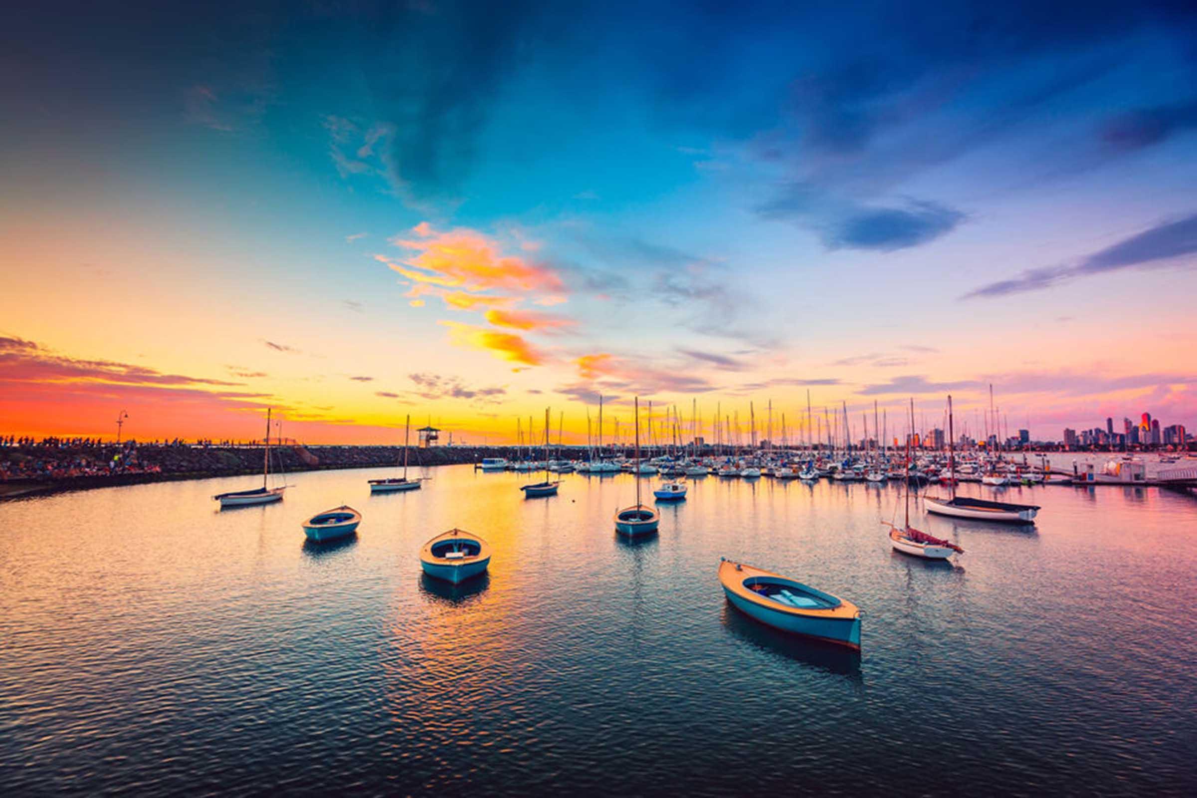 Colorful sunset over a peaceful marina with sailboats anchored on calm water and a city skyline in the distance.