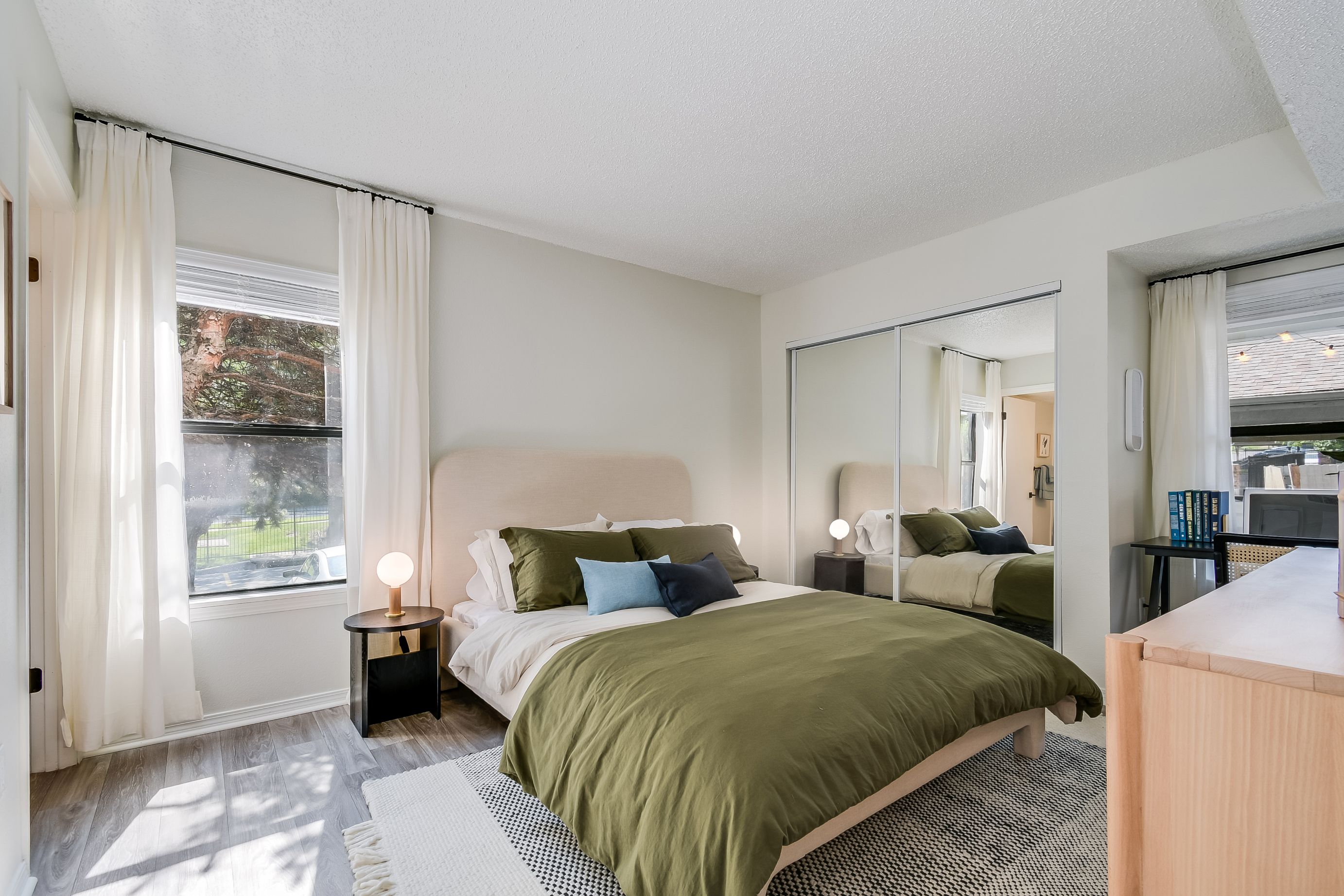 Photograph of a modern bedroom featuring a queen-sized bed with olive green bedding, multiple pillows, and a beige upholstered headboard. Room includes two windows with white curtains, a black nightstand with a round lamp, mirrored closet doors, a light wood dresser, and a patterned area rug on light hardwood floors.