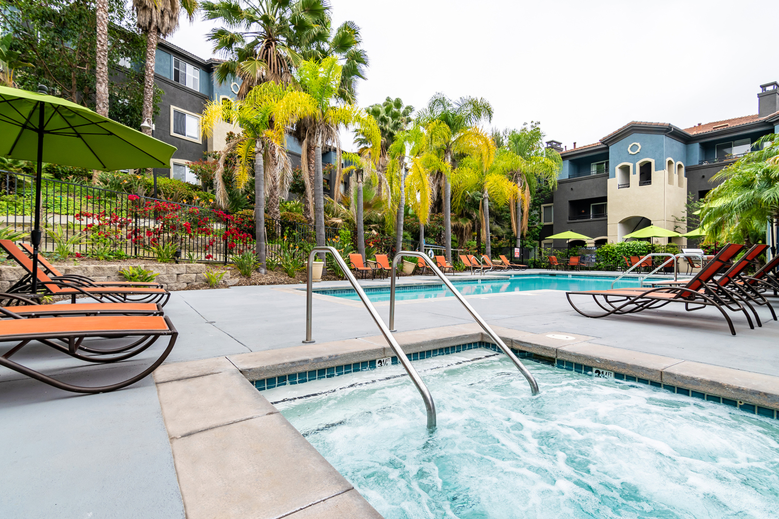 Pool area viewed from spa hot tub with lounge chairs, palm trees, and a building exterior in the background