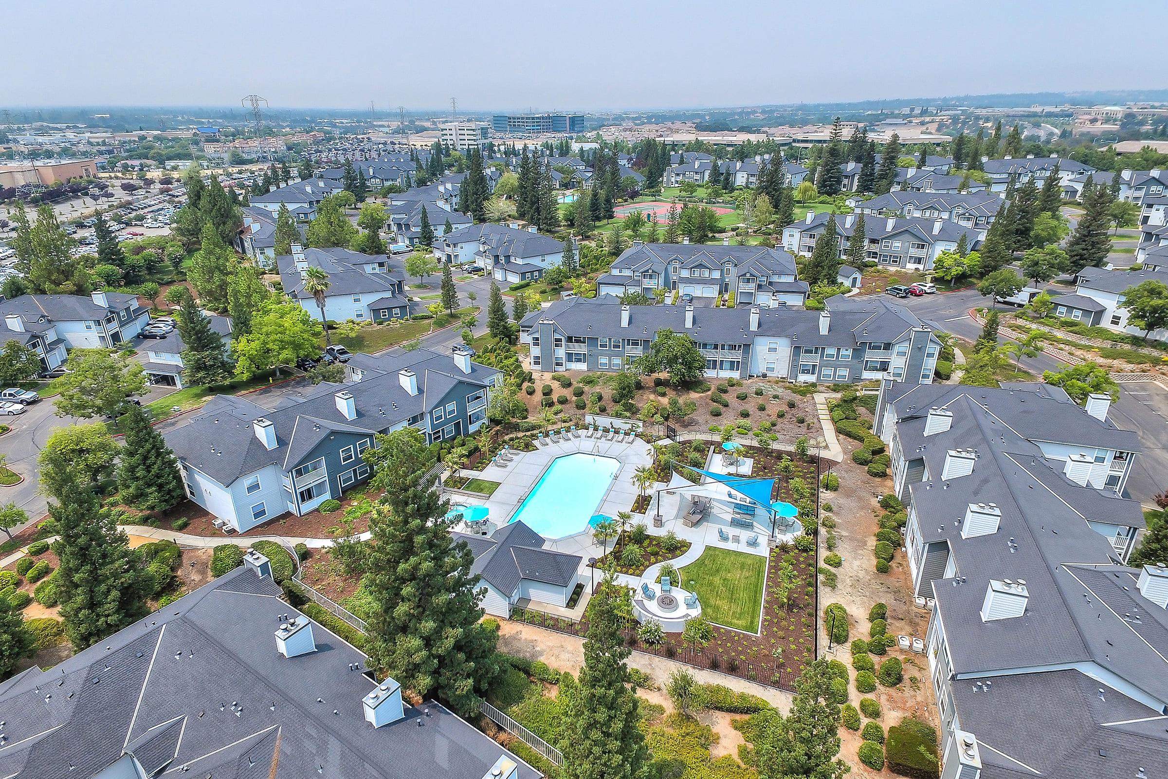 Aerial view of a landscaped apartment community featuring a central outdoor swimming pool, shaded lounge areas, fire pit gathering space, walking paths, mature trees, and surrounding residential buildings.