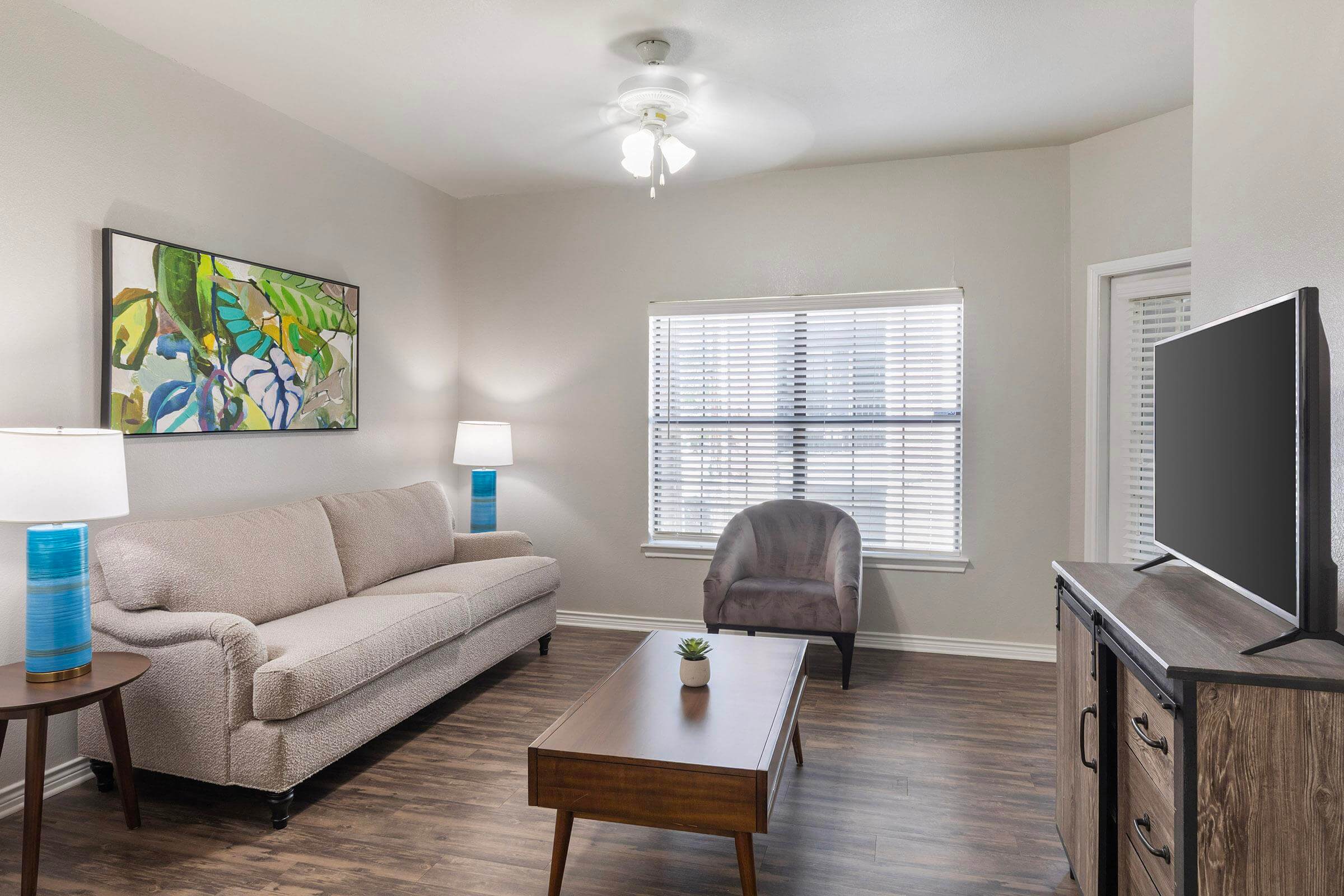 living room with wood flooring and a ceiling fan, a couch, side tables with blue lamps, coffee table, upholstered chair in front of a large window with blinds, and a tv on a stand.
