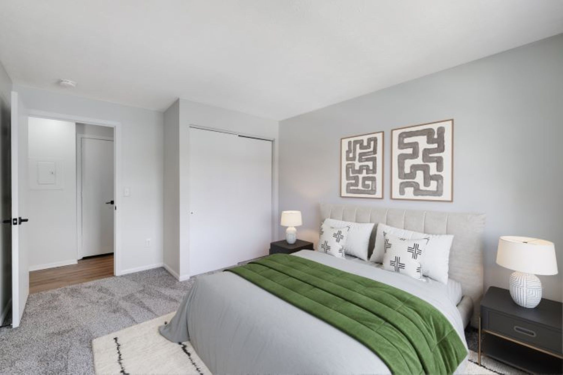 Photograph of a modern bedroom featuring a neatly made bed with gray and green bedding, two black nightstands with white lamps, and two abstract framed artworks above the headboard. Room has light gray walls, carpeted floor, a white closet door, and an open doorway leading to a hallway.
