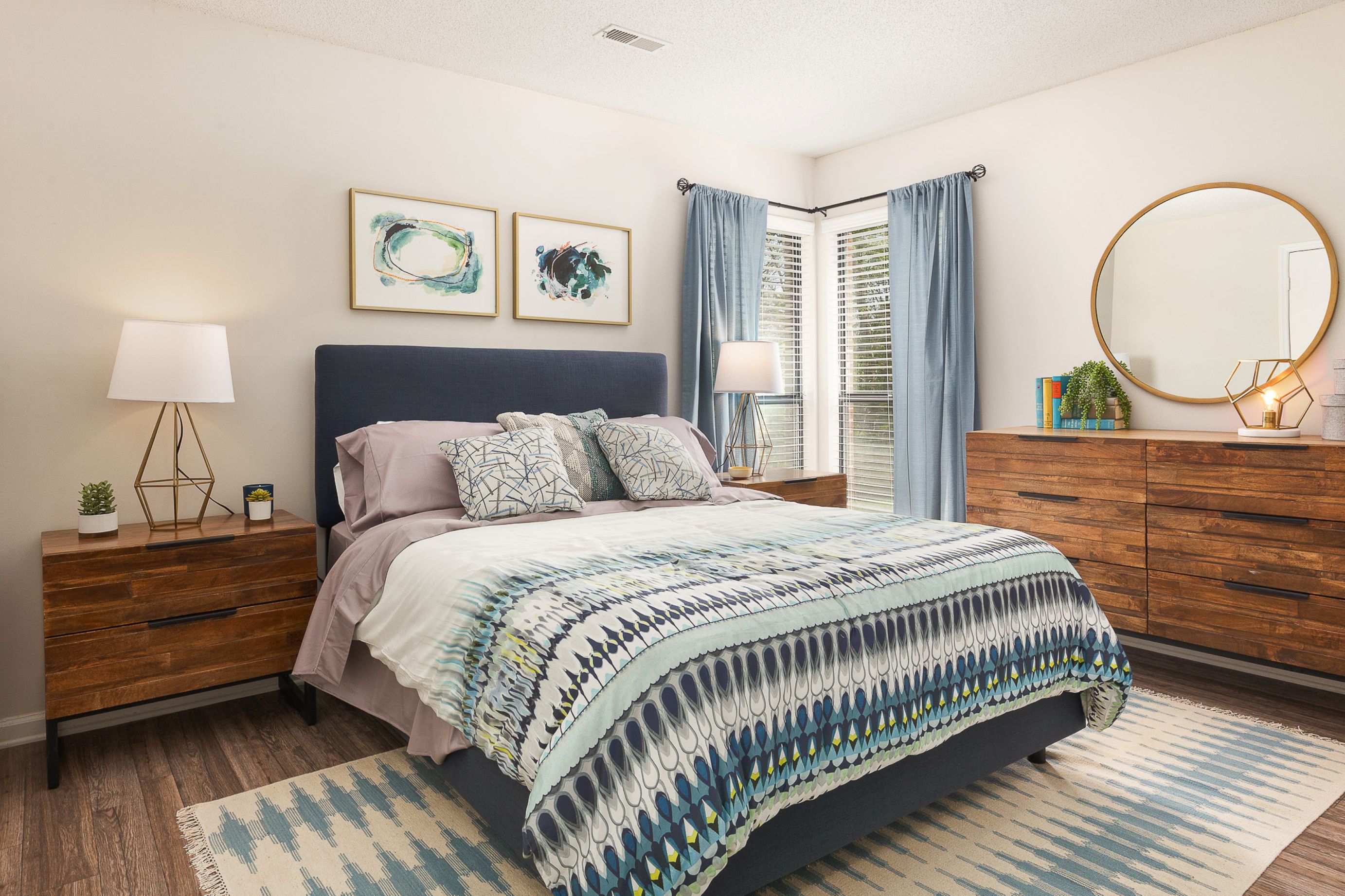 Photograph of a modern bedroom featuring a queen-sized bed with patterned blue and white bedding, flanked by wooden nightstands with lamps and decorative plants. The room includes a wooden dresser with a round mirror, blue curtains on corner windows, and abstract artwork above the bed, creating a cozy and stylish atmosphere.
