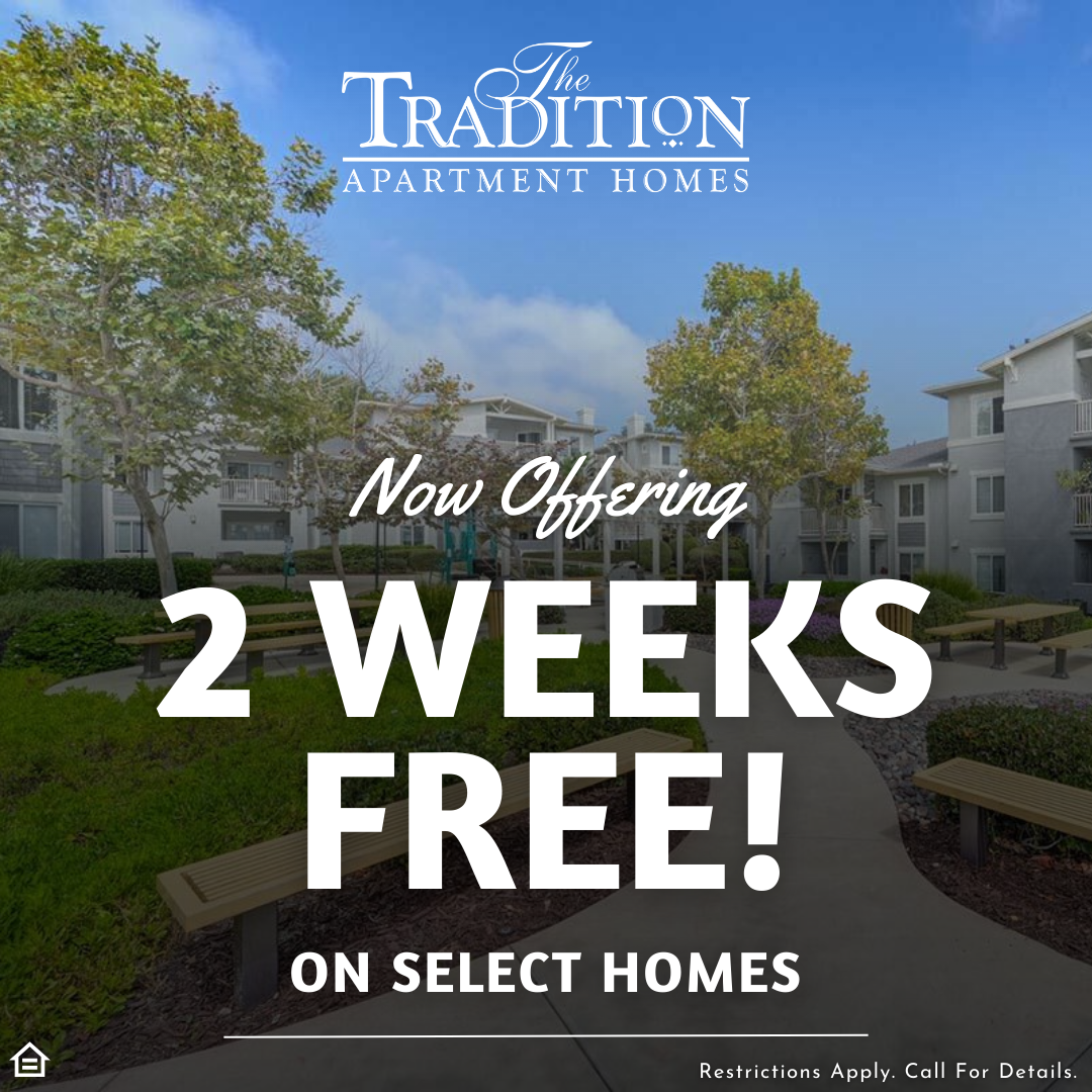 An image of a promotional graphic overlaid on an exterior photo of an apartment courtyard. The graphic features the logo for "The Tradition Apartment Homes" at the top center. In the center, there is large white text that reads "Now Offering 2 WEEKS FREE!", with smaller text below stating "ON SELECT HOMES". The background photo shows two grey apartment buildings with balconies and extensive green landscaping, including trees, bushes, a concrete pathway, and multiple wooden benches under a twilight sky with