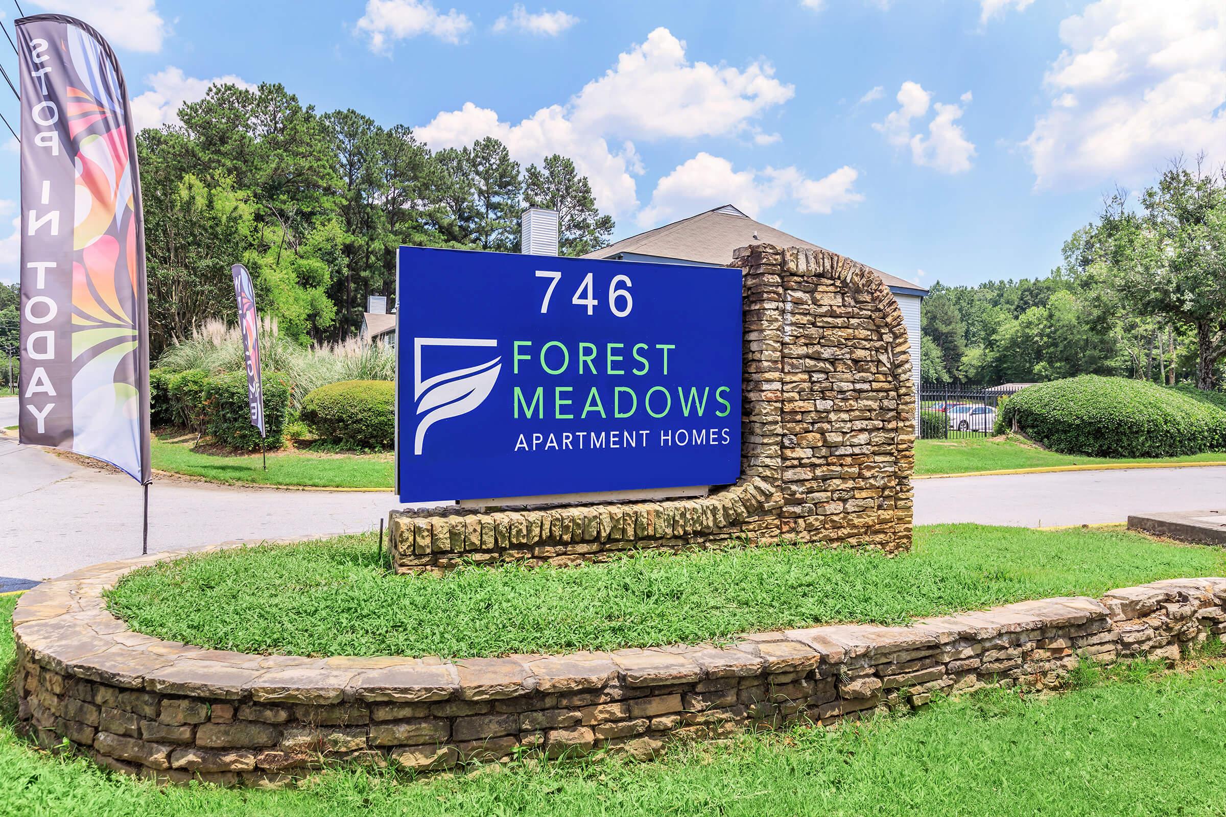 Sign for Forest Meadows Apartment Homes displaying the address "746," surrounded by lush greenery and a stone wall. There are blue skies with a few clouds in the background, and colorful flags that promote leasing opportunities.