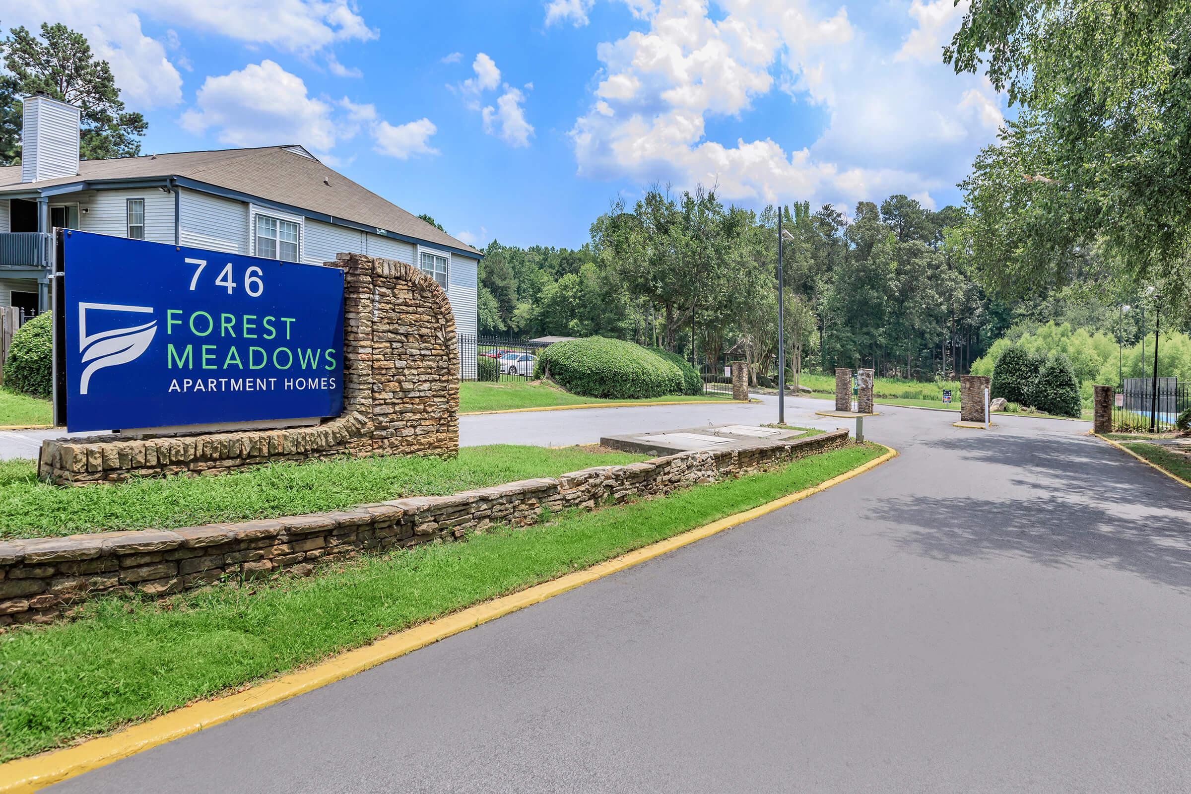 Sign for Forest Meadows Apartment Homes located at 746. The entrance features landscaped greenery, stone accents, and a clear blue sky in the background. A smooth road leads into the complex, flanked by manicured bushes and trees.