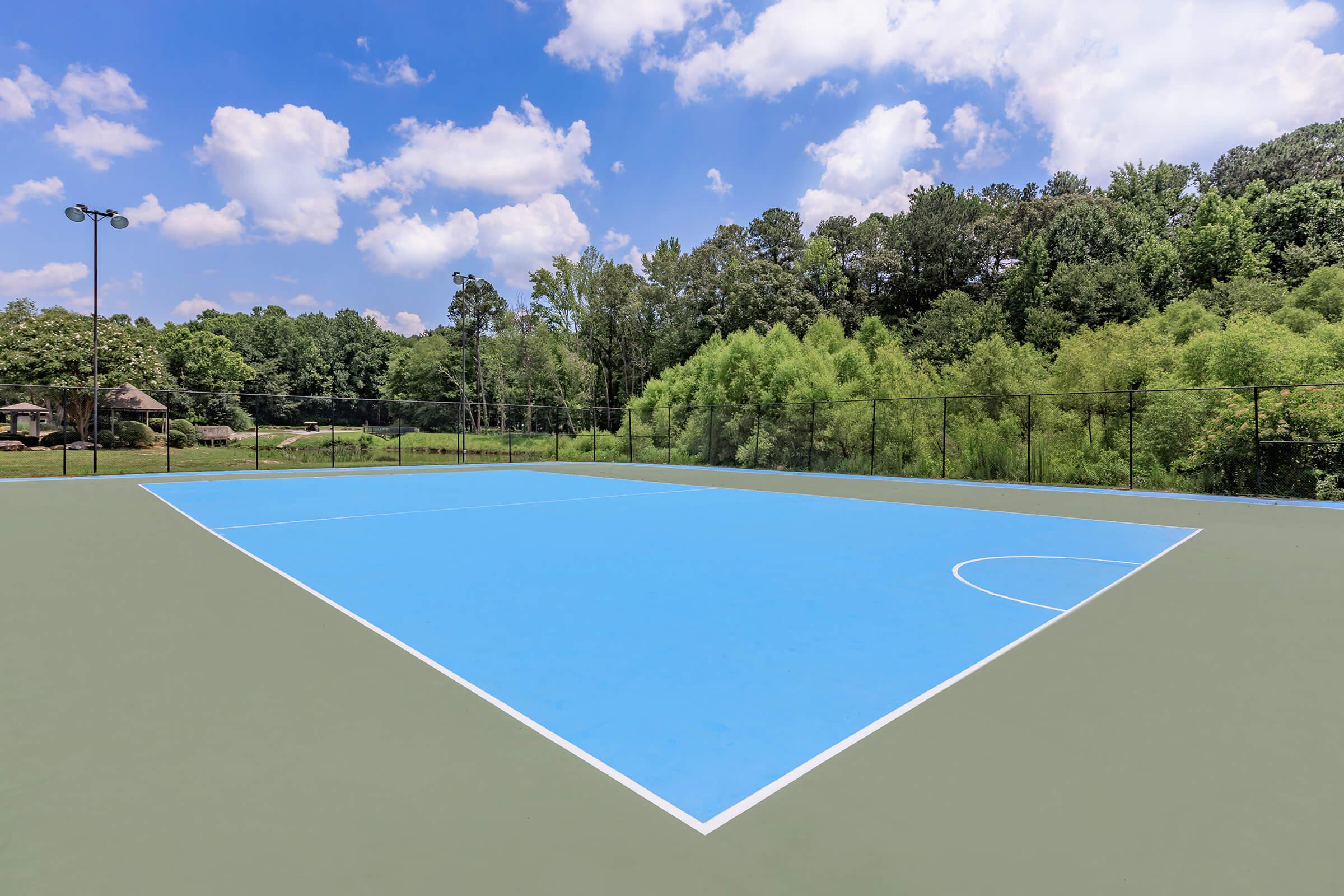 A bright blue sports court surrounded by a green landscape, with trees in the background and a partly cloudy sky above. The court has white lines marking the boundaries and is situated within a fenced area. Ideal for outdoor activities and games.
