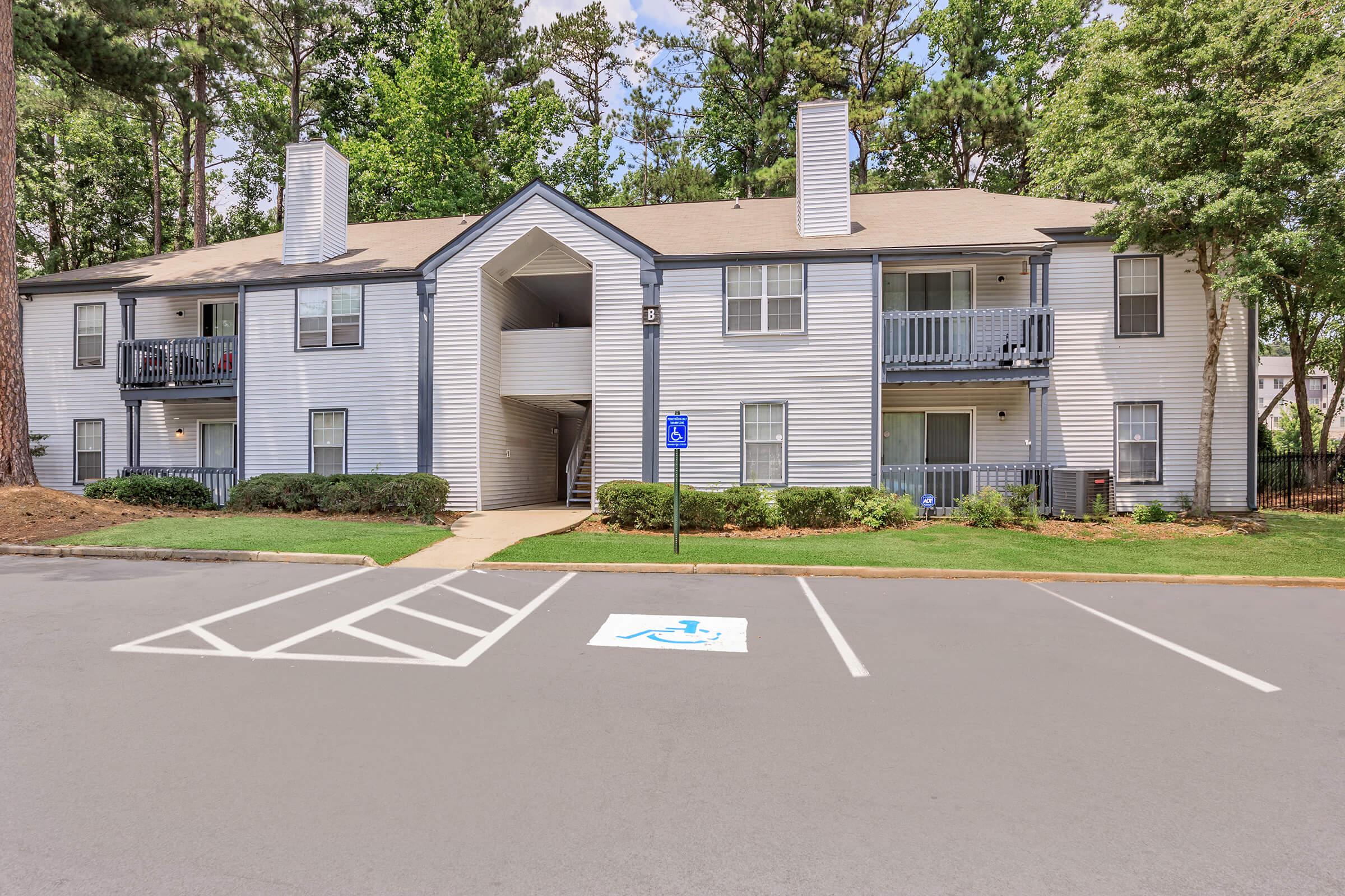 A two-story apartment building with light gray siding, featuring several balconies. The building is surrounded by trees and landscaped grass. A designated handicapped parking space is visible in front of the building, with a paved area leading to the entrance.