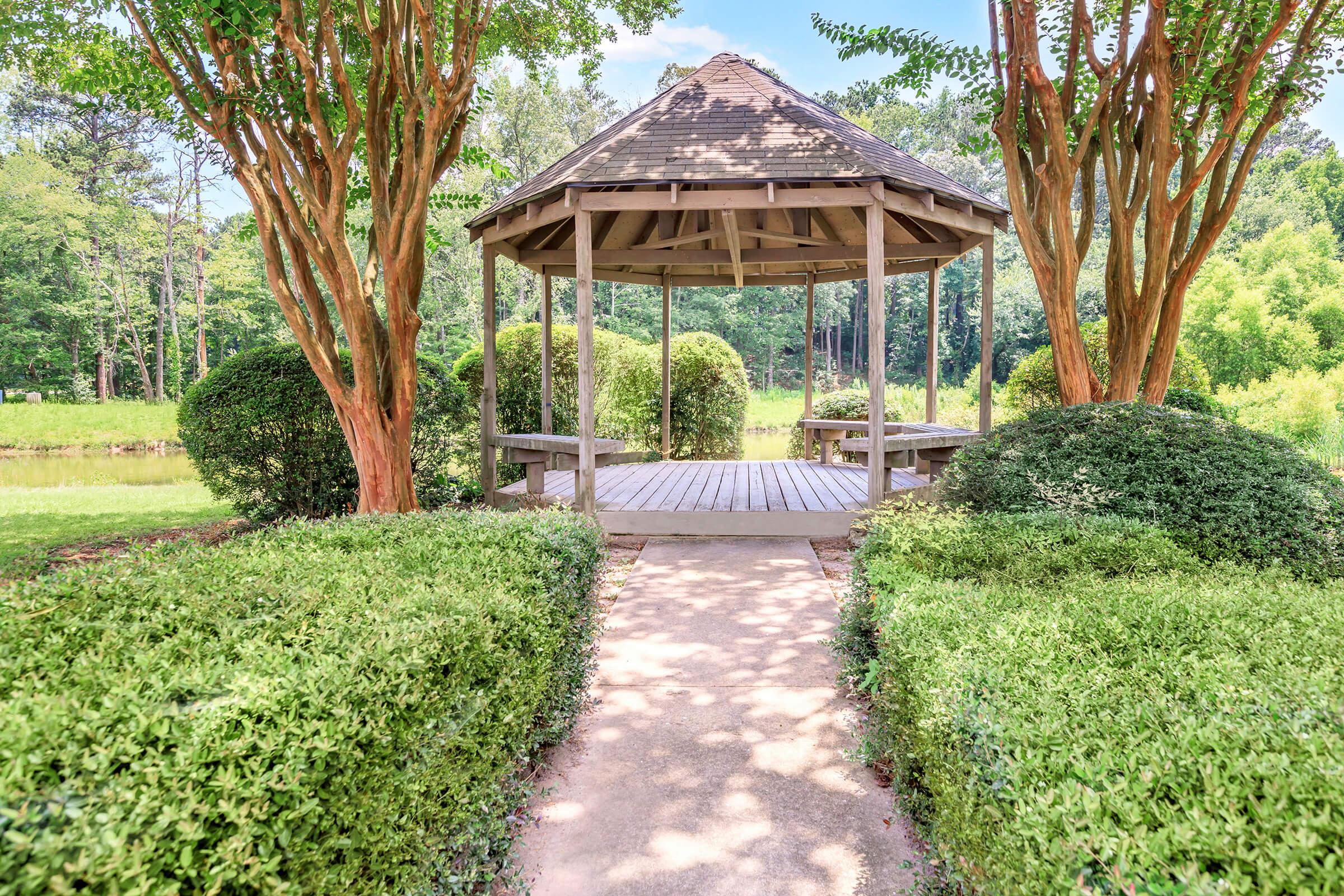 A peaceful gazebo surrounded by lush greenery and trimmed bushes, with a stone pathway leading towards it. The scene features dappled sunlight filtering through the trees, creating a serene outdoor atmosphere in a park-like setting.