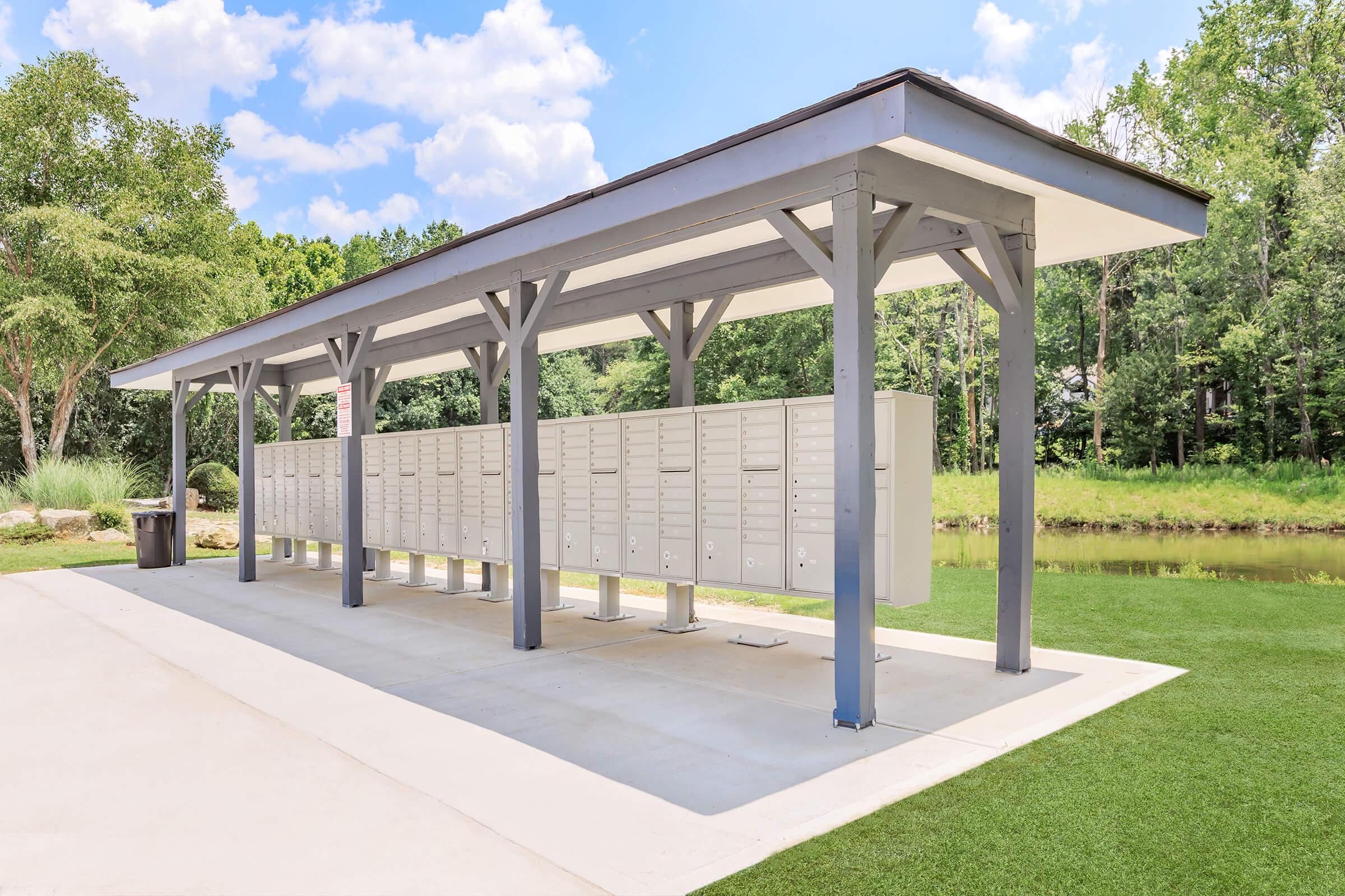 A row of outdoor mailboxes under a covered structure, set in a landscaped area with green grass and trees in the background. The sky is partly cloudy, and there is a small pond nearby, creating a peaceful and inviting atmosphere for mail collection.