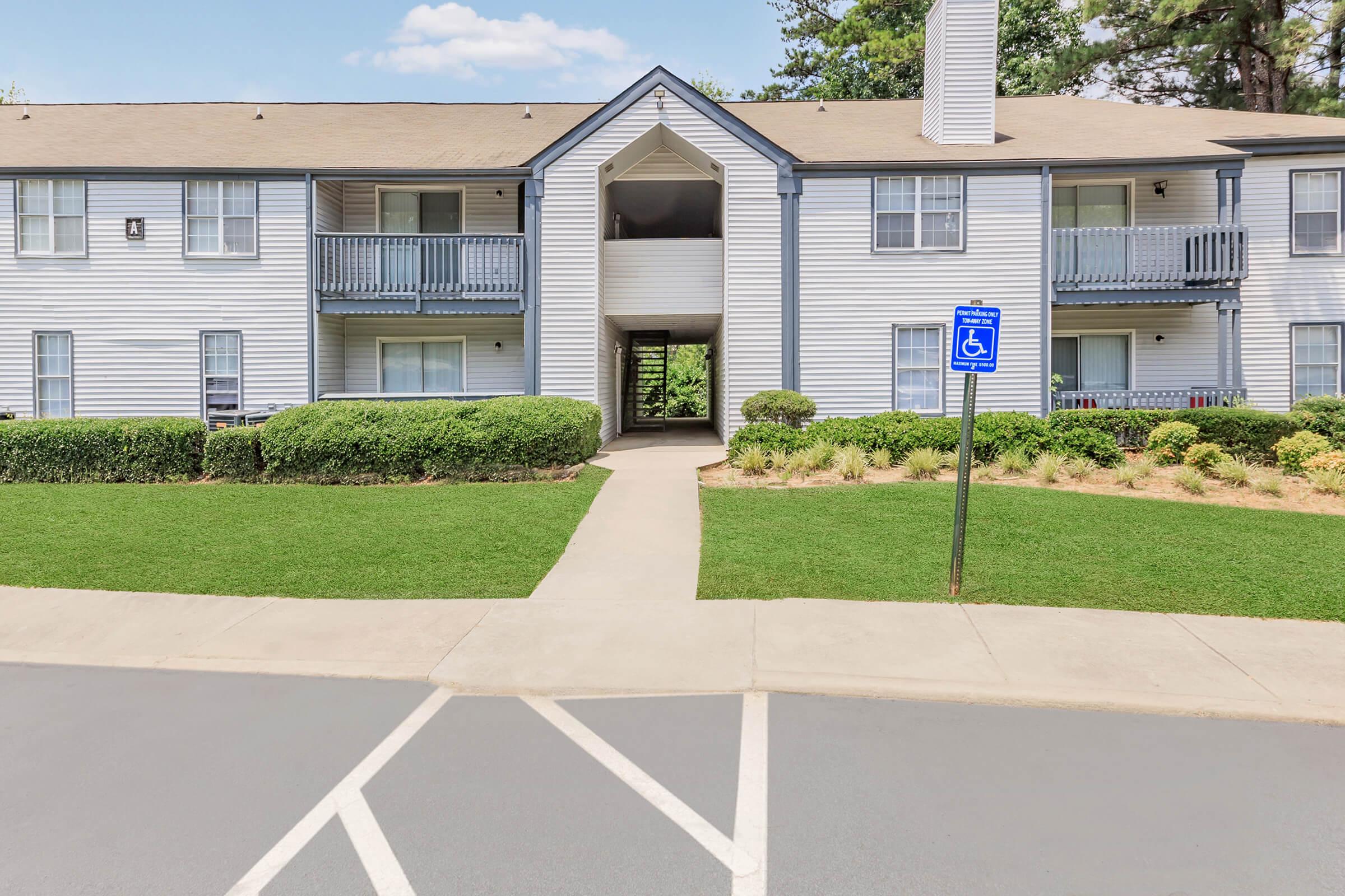 A view of a two-story residential building with white siding and blue accents, featuring balconies and an entrance between the two wings. The front grounds are landscaped with neatly trimmed bushes and grass, and a designated accessible parking sign is visible in the foreground.