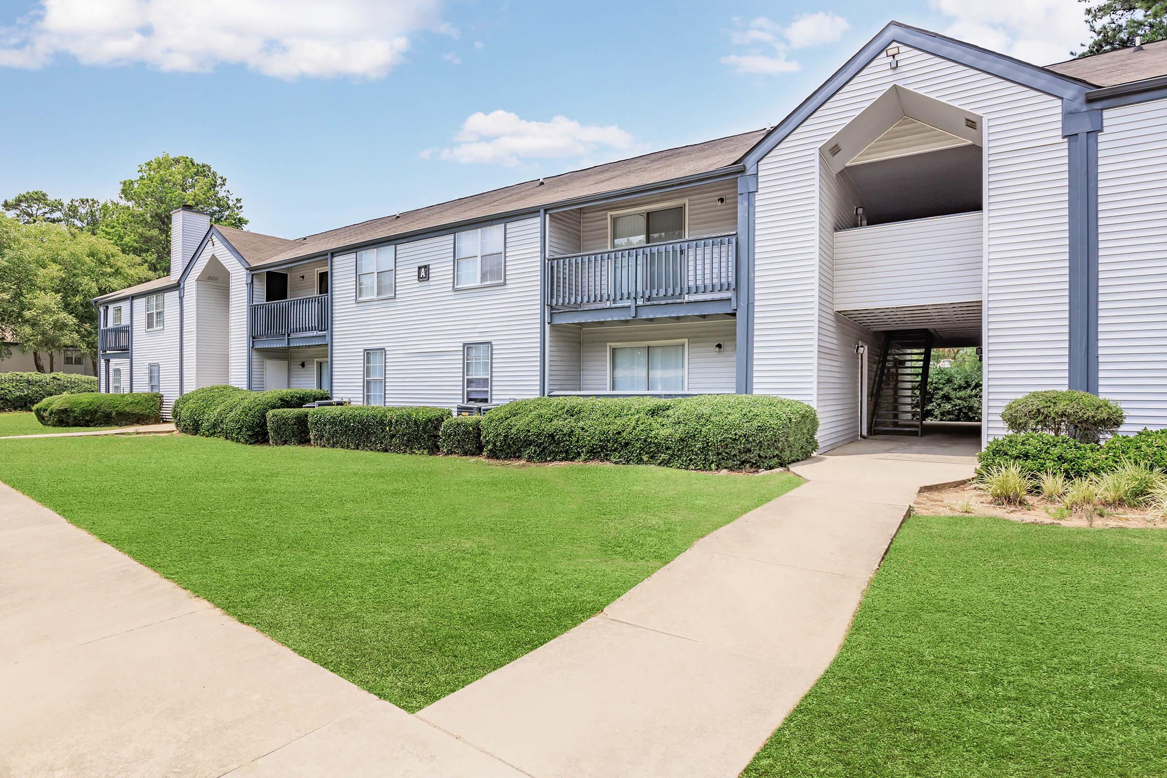A view of a well-maintained apartment complex featuring two-story buildings with blue accents. Lush green lawns and neatly trimmed bushes surround the pathway leading to the entrance. Clear blue skies and fluffy clouds enhance the inviting atmosphere of the community.