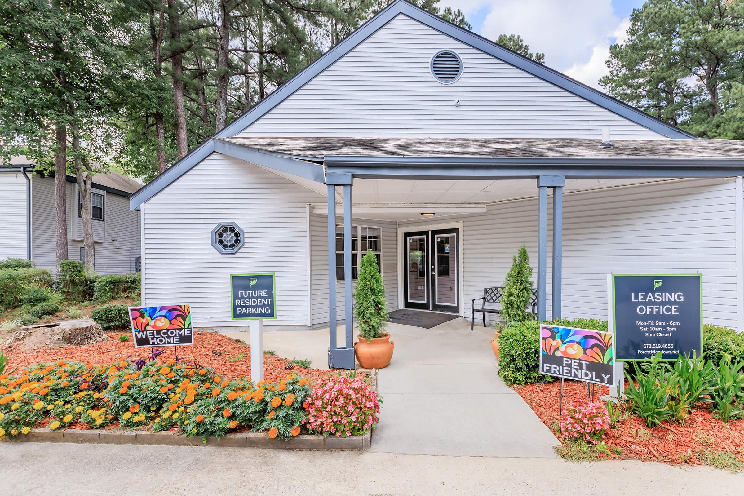 Leasing office exterior of an apartment complex, featuring a welcoming entrance with a sign that says "Welcome Home," a "Pet Friendly" sign, and designated parking for future residents. The area is landscaped with colorful flowers and surrounded by trees.