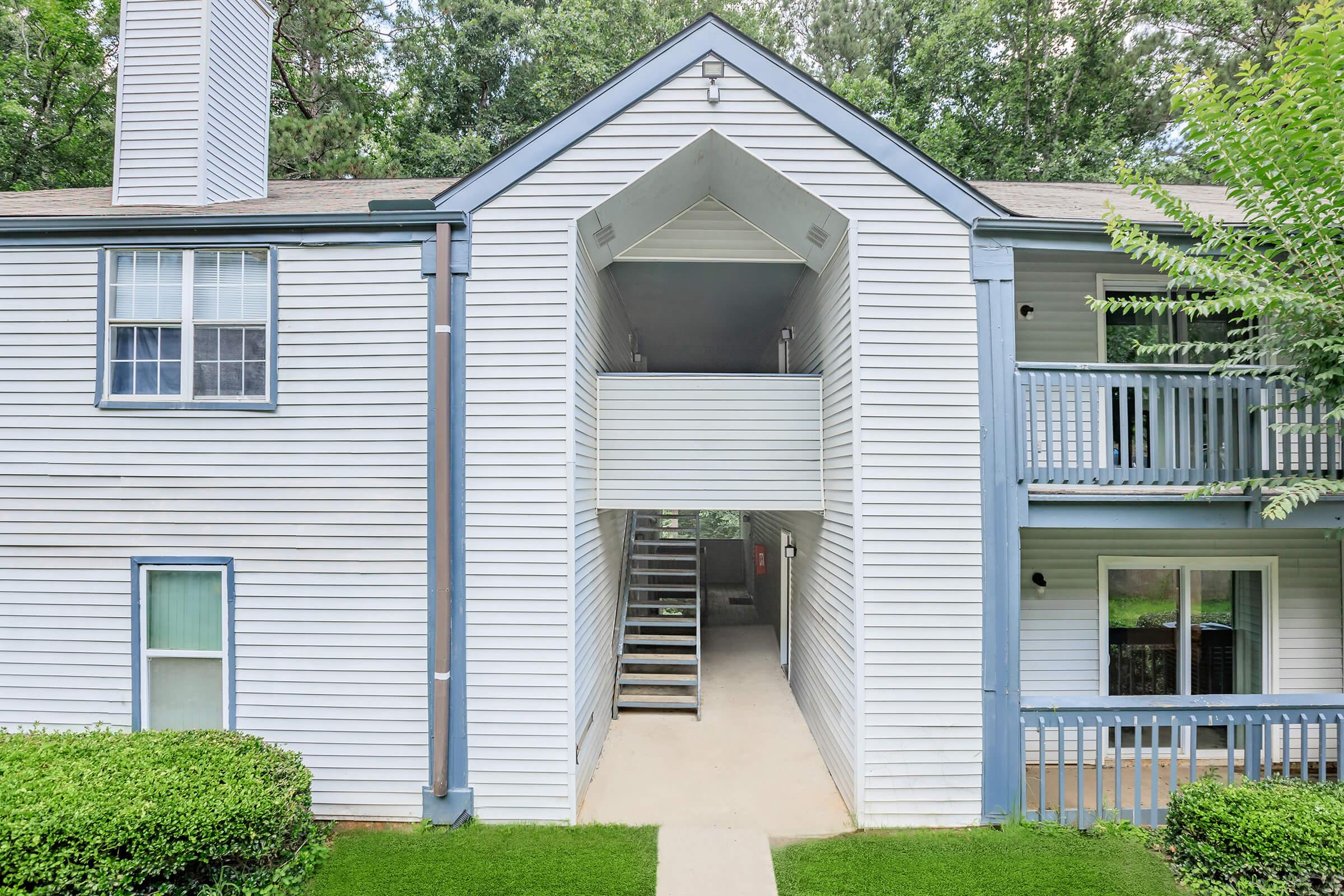 View of a multi-unit apartment building featuring white siding, a central entrance with stairs leading to the upper level, and surrounding greenery. The exterior includes balconies and well-maintained landscaping, creating an inviting residential atmosphere.