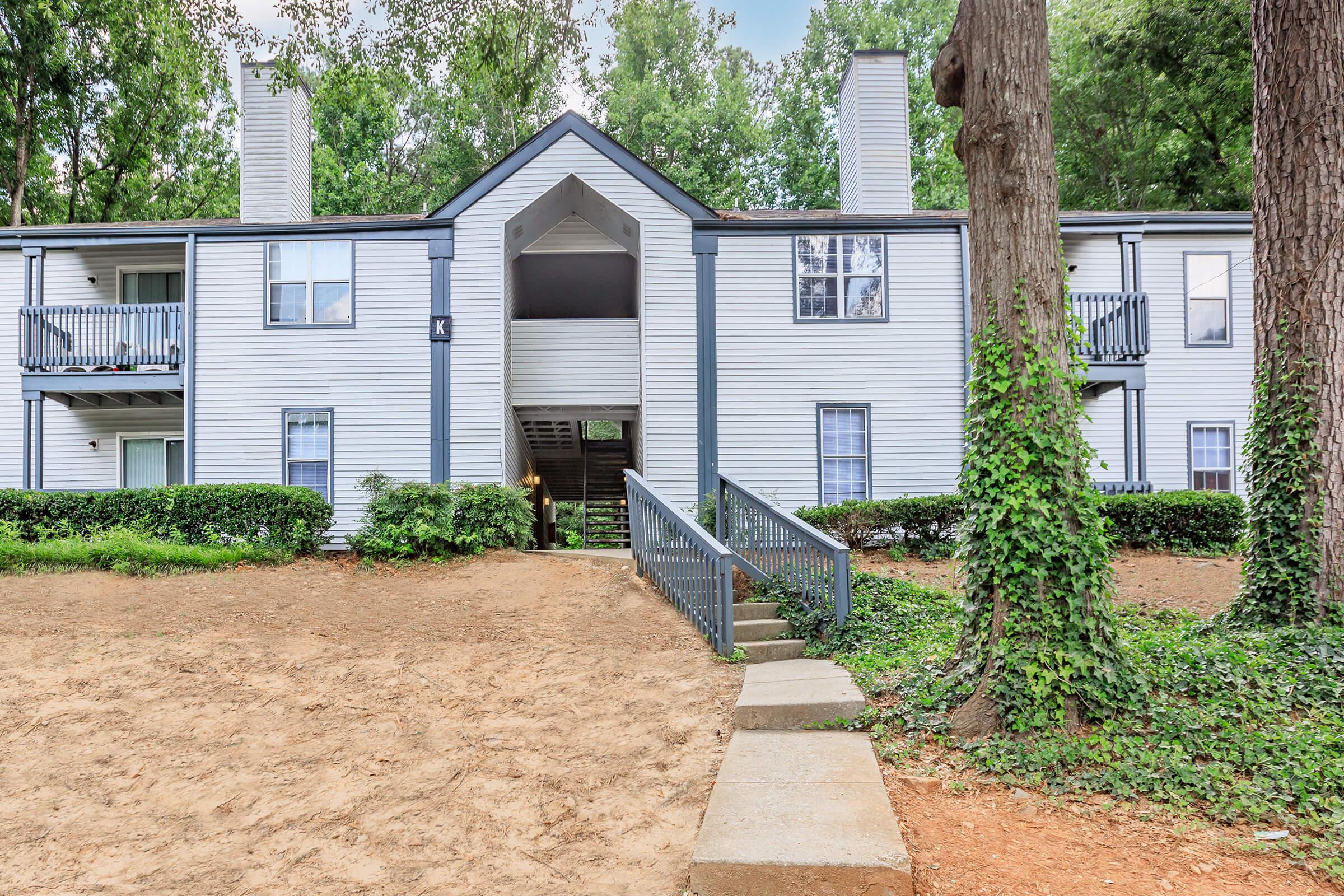 A two-story apartment building with a light-colored exterior and dark trim, surrounded by trees and greenery. The entrance features a pathway leading up to a central archway, flanked by balconies on the upper level. The landscape includes a sandy area and landscaped shrubs.