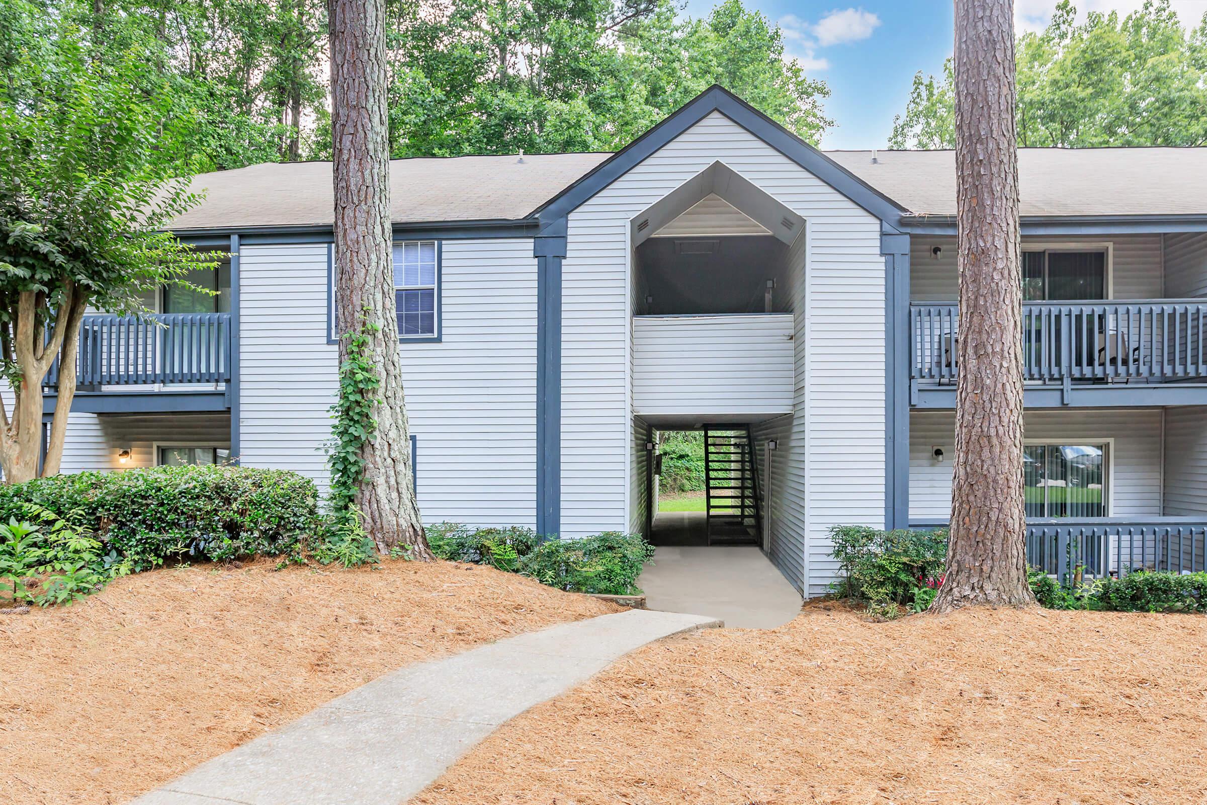 Exterior view of a multi-unit residential building featuring a light blue and white facade. Two tall pine trees flank the entrance, which has a concrete pathway leading to an enclosed entryway. The surrounding area has well-maintained landscaping with shrubs and mulch.