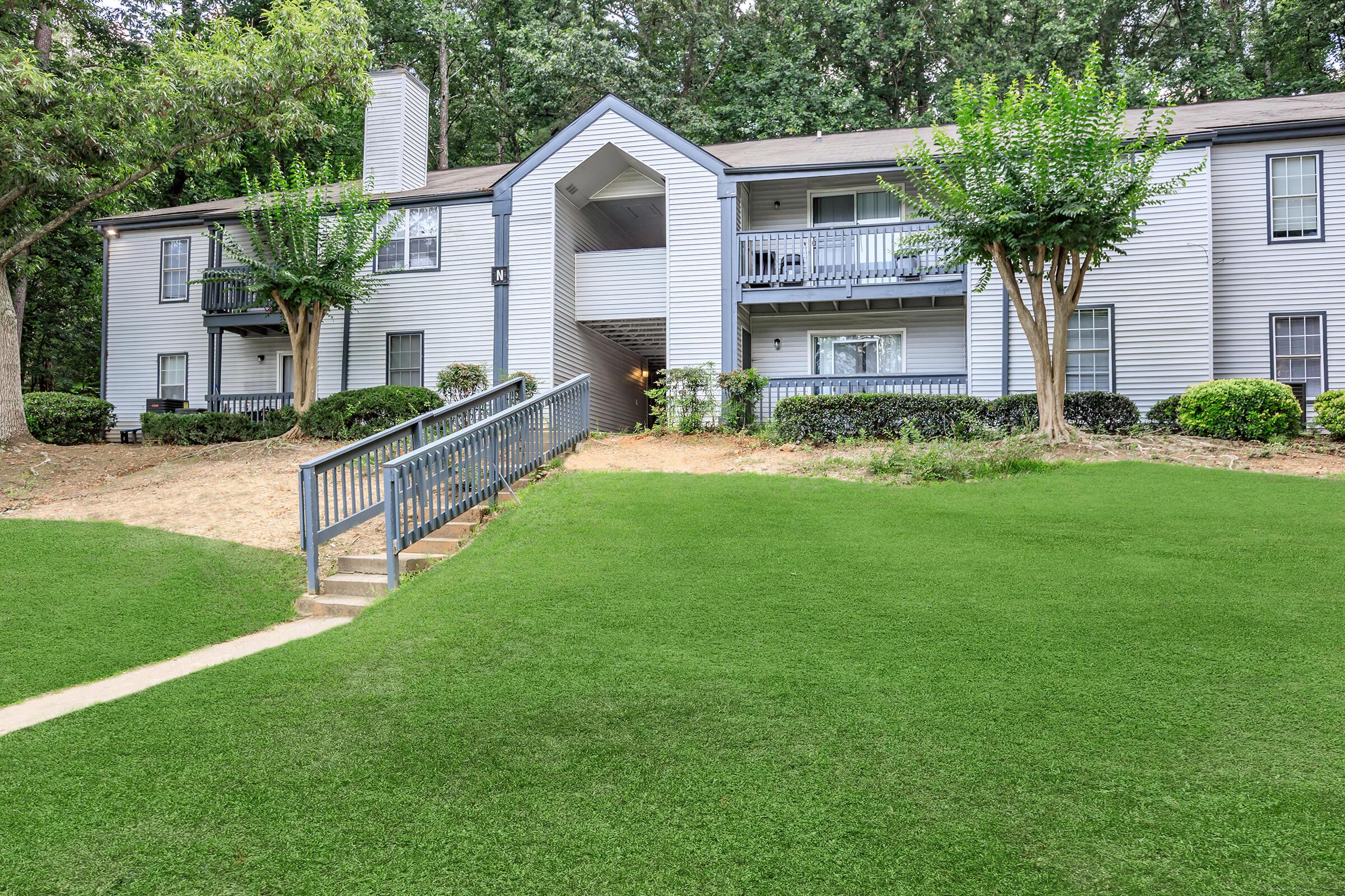 A multi-unit apartment building surrounded by green lawns and trees. The building features a light gray exterior with balconies and a staircase leading to an entrance. Lush greenery and landscaping enhance the peaceful, residential atmosphere.
