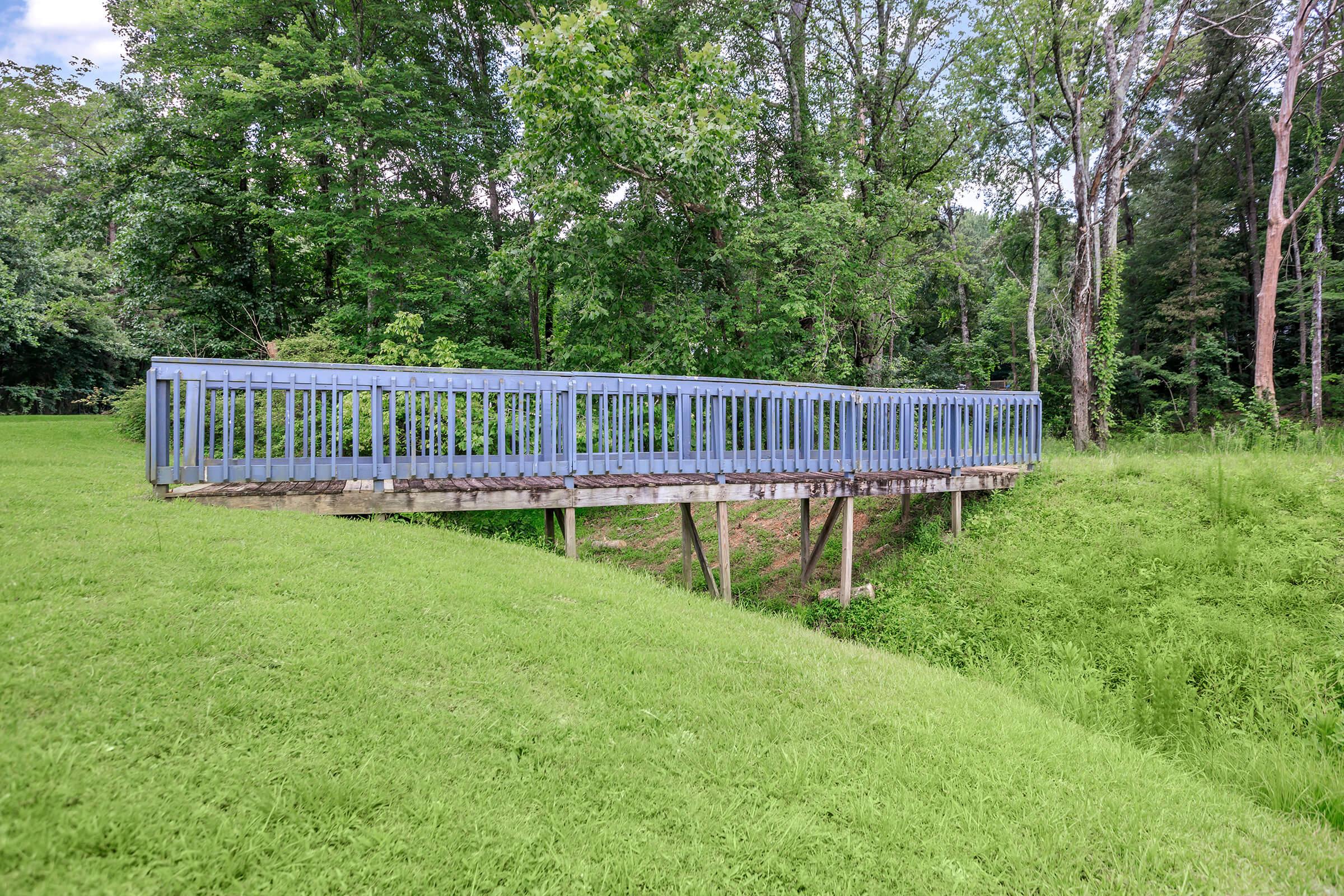 A blue wooden footbridge spans a small creek, supported by stilts above a grassy area. Surrounding the bridge, tall green grass and dense trees create a serene natural setting under a clear sky.