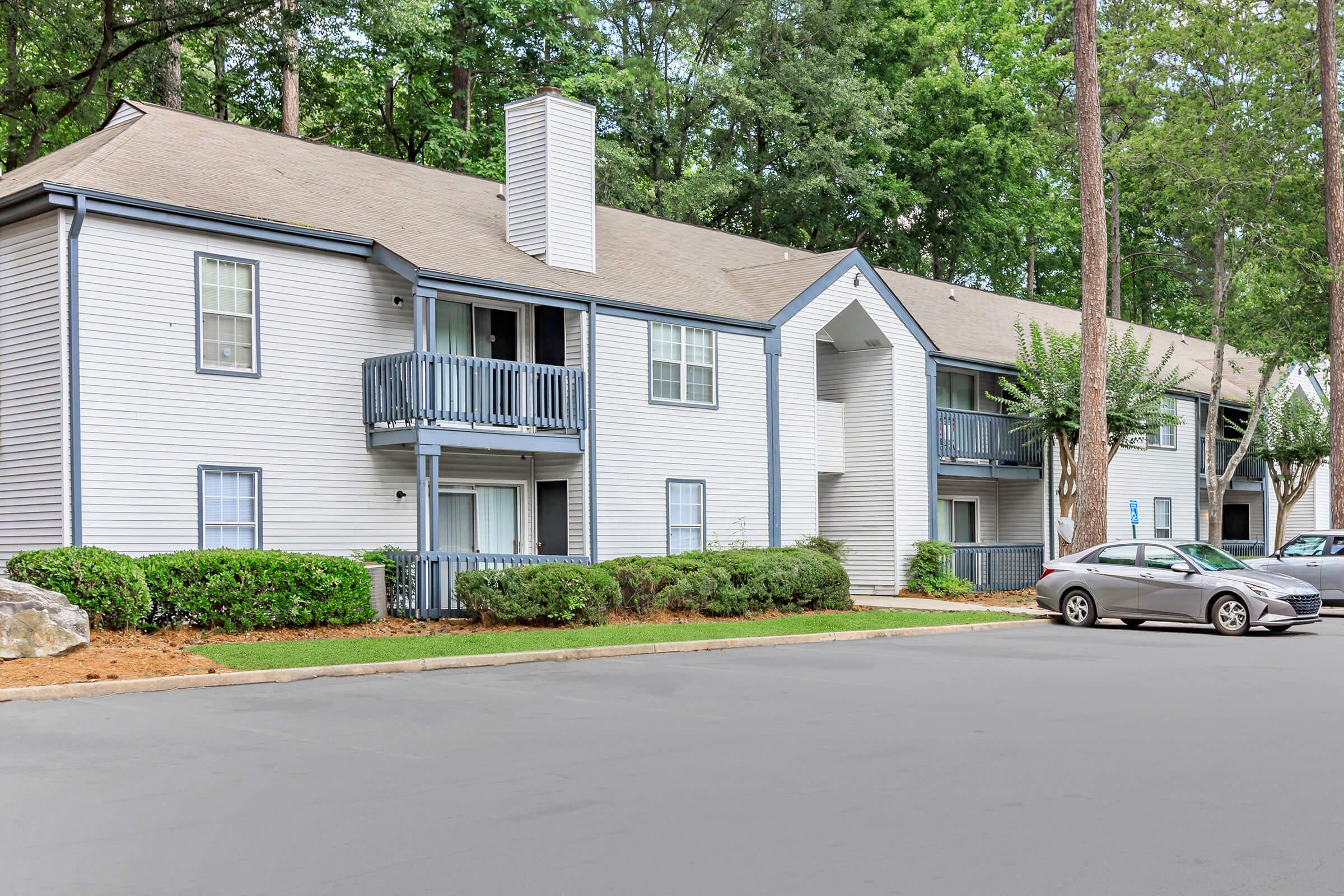 Two-story apartment building with multiple units featuring blue trim and balconies. The exterior has light-colored siding surrounded by green landscaping and trees. A parked car is visible in the foreground on a paved driveway.