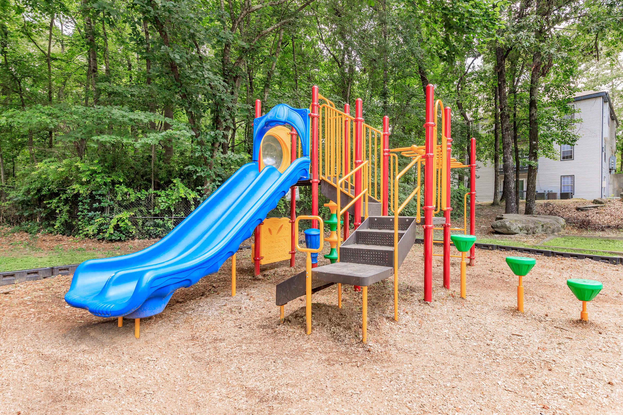 Colorful children's playground featuring a blue slide, climbing structure, and various play features. The area is surrounded by greenery and has gravel flooring for safety. A few green play funnels are positioned around the base, and residential buildings are visible in the background.