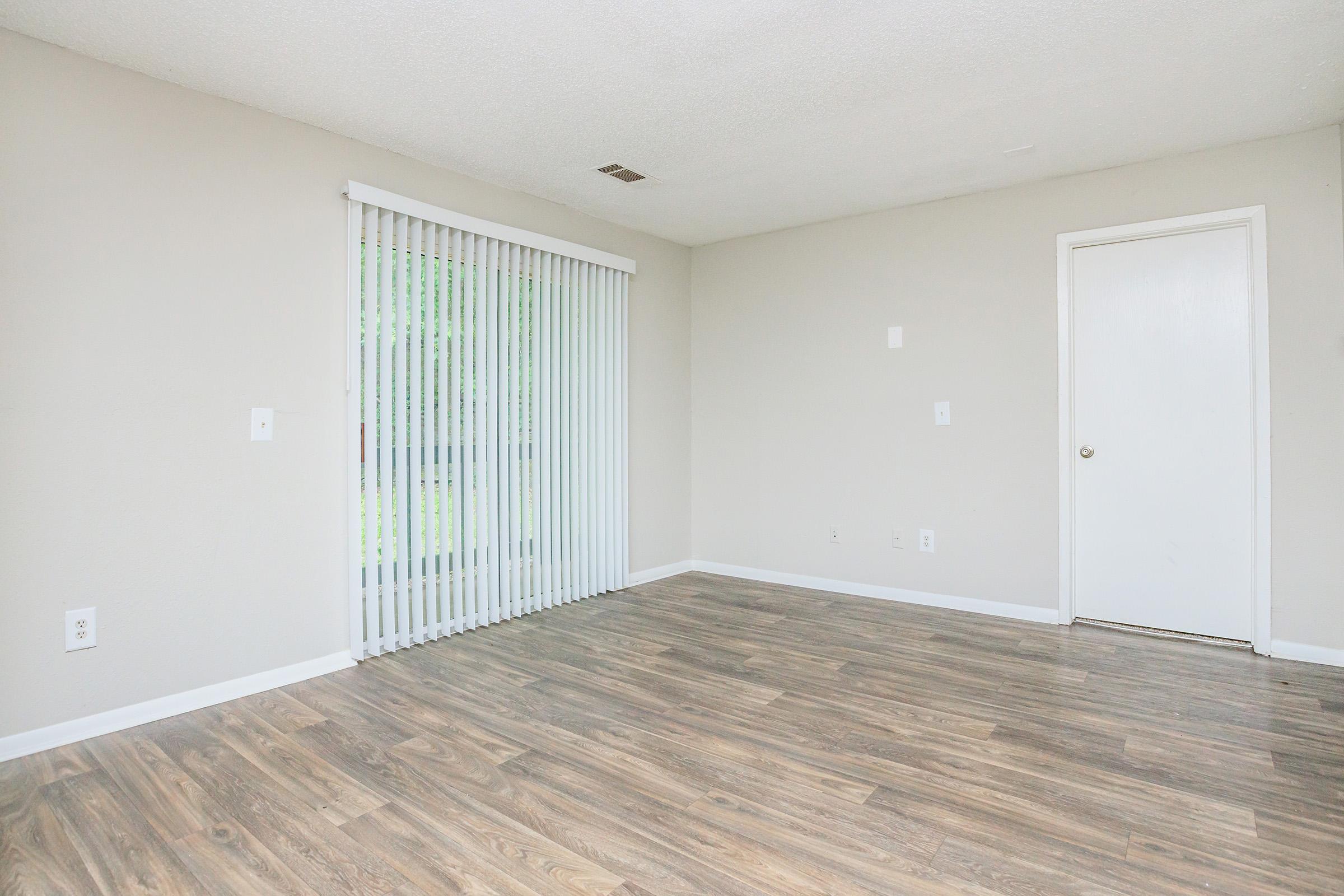 A vacant room with light brown wood-style flooring, neutral-colored walls, and a sliding glass door with vertical blinds leading to an outdoor area. A plain door is visible on the right side. The space is well-lit and clean, suitable for design or decoration.
