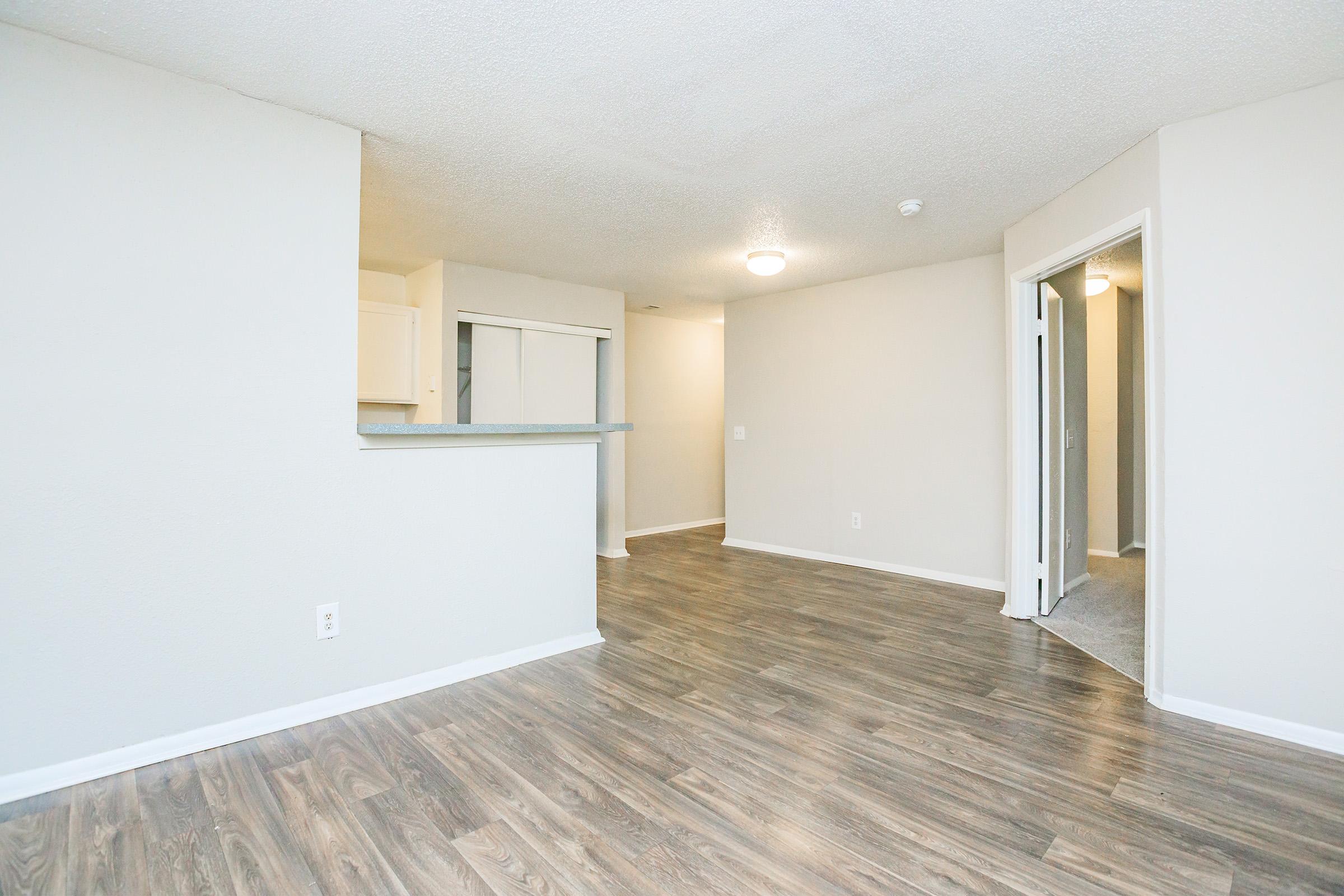 A spacious, well-lit room with light-colored walls and modern wood-like flooring. It features an open layout with a small partition to a kitchen area, a doorway leading to another room, and ample natural light from overhead lighting. The overall look is clean and inviting, suitable for a living space.