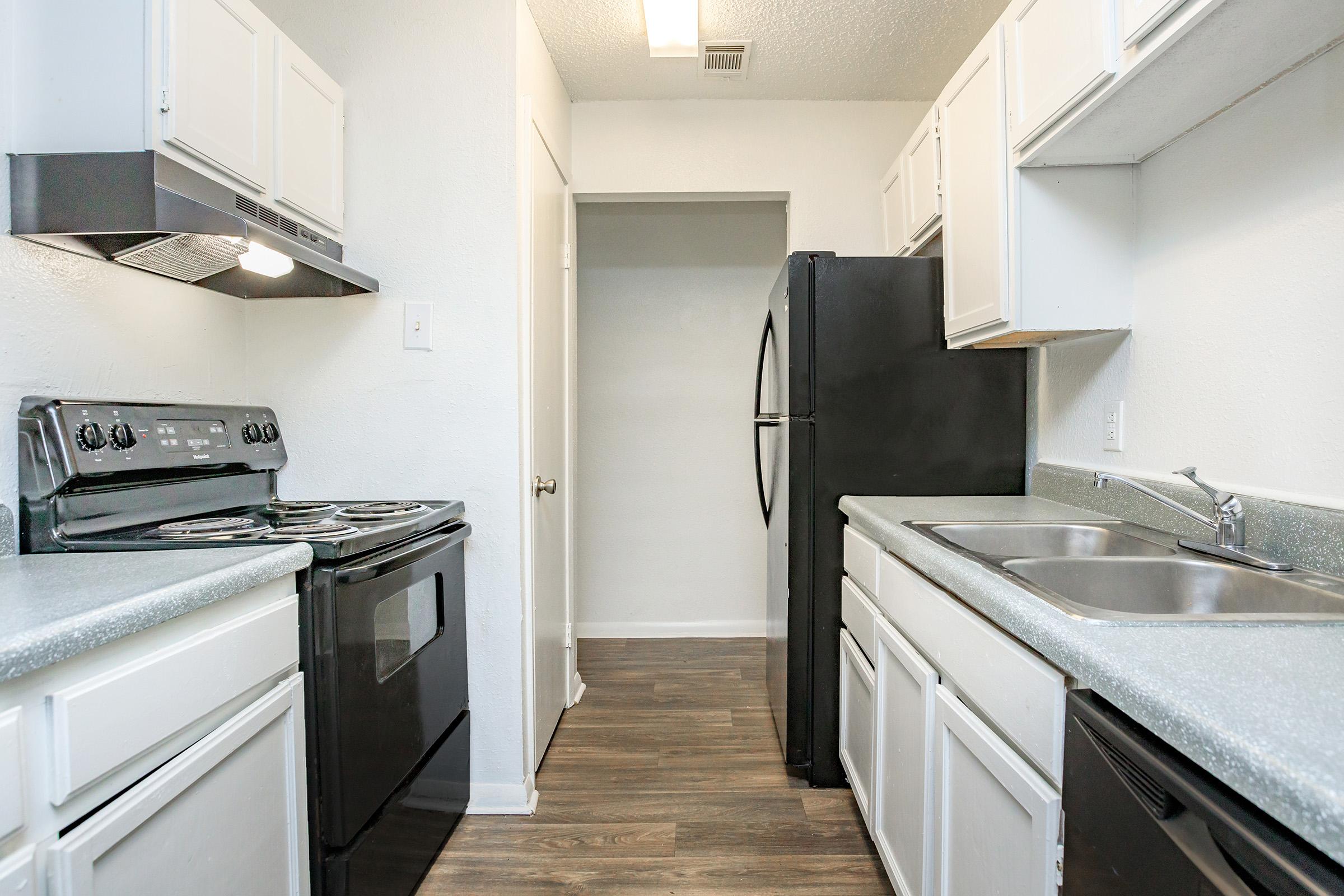 Modern kitchen featuring white cabinetry, a black refrigerator, and black appliances, including a stove and dishwasher. The countertops are gray, and the flooring is laminate. The space appears bright and well-lit with natural light.