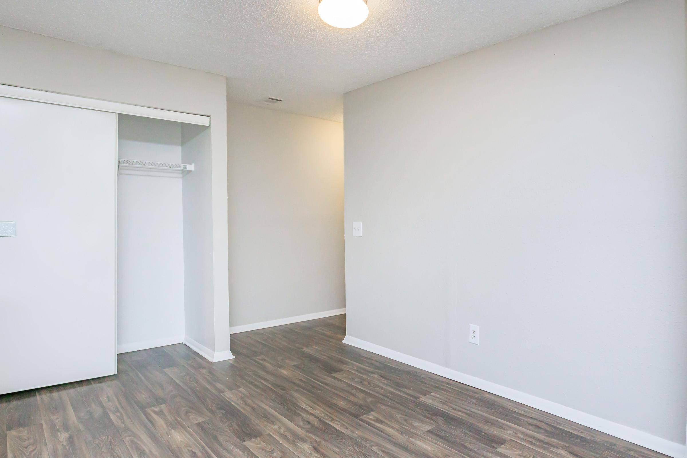 A clean and empty interior space featuring light gray walls, a wooden floor, and a ceiling light. On one side, there is an open closet with sliding doors, while the rest of the room is unadorned, offering a blank canvas for decoration or furnishing.
