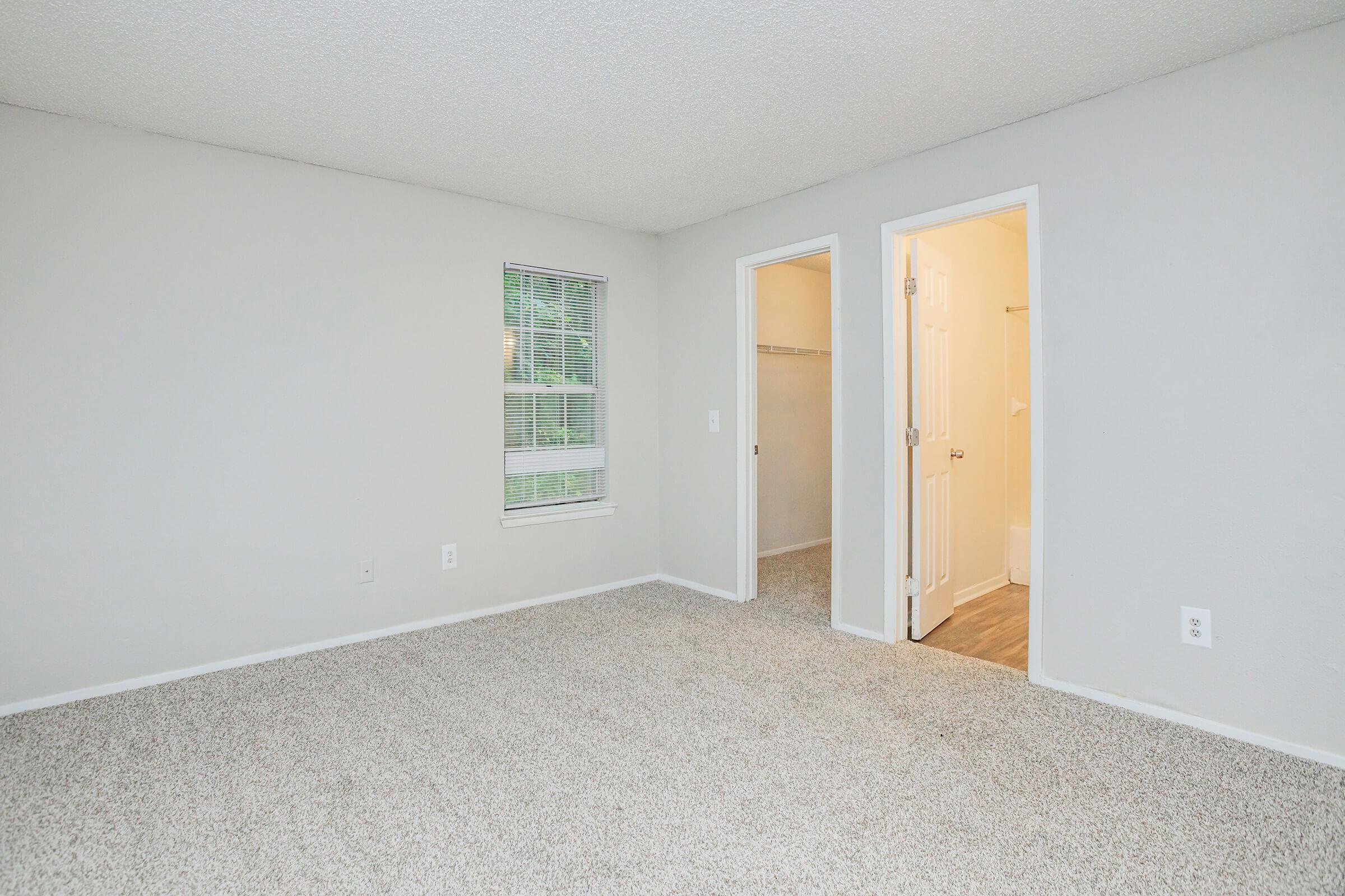A spacious, empty room with light gray walls and carpeted flooring. It features a window with blinds, a small closet entrance, and an open door leading to a bathroom. The overall atmosphere is bright and inviting, with natural light filtering in.