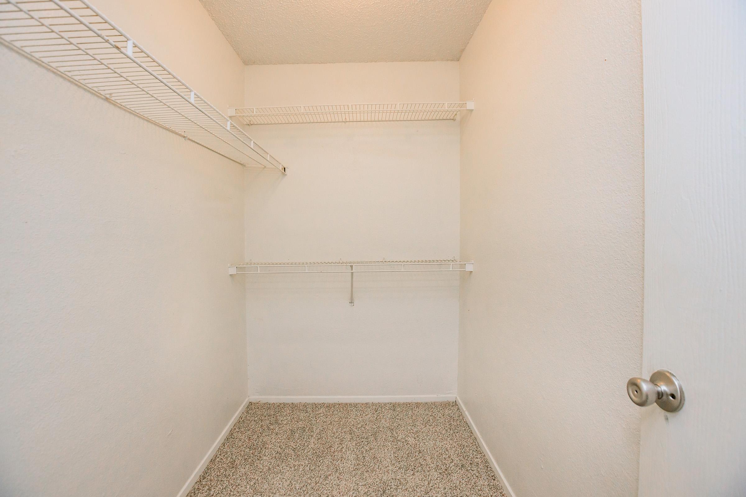 Empty closet with white walls, a light-colored carpeted floor, and two wire shelves mounted on the walls. The door handle is visible on the right side. The space appears clean and well-lit, but lacks any items or storage within.