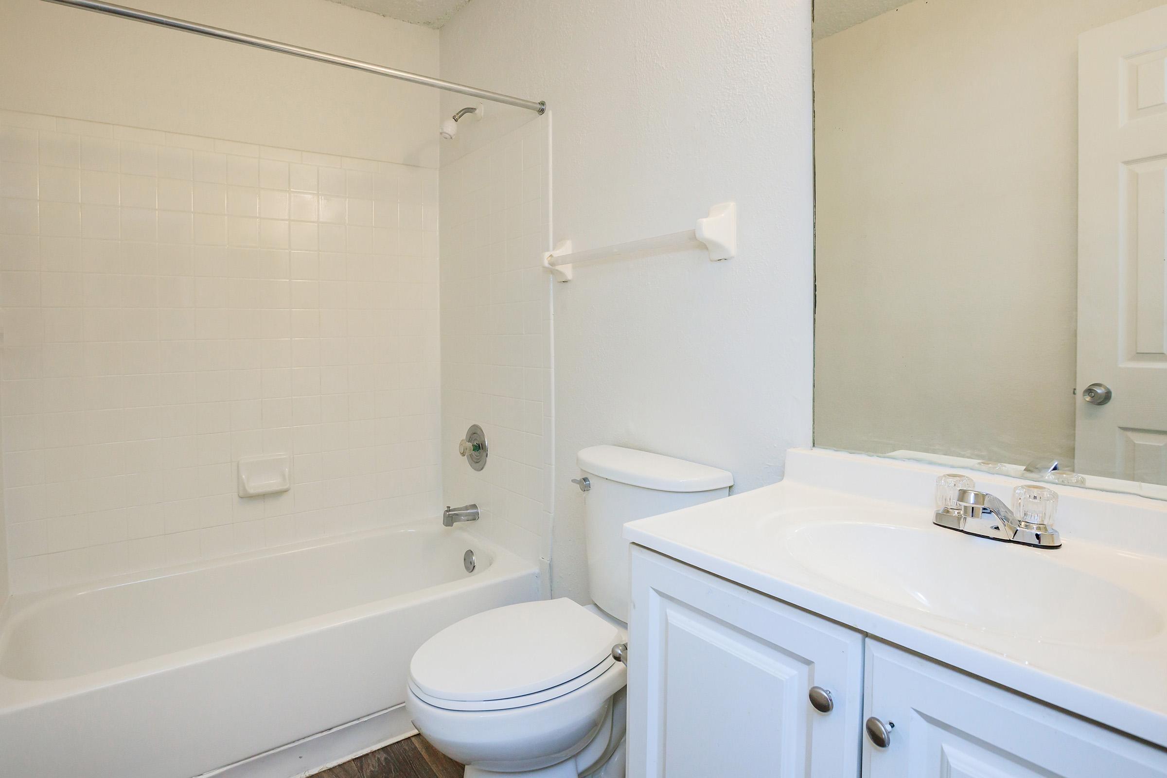 A clean, modern bathroom featuring a white bathtub with a shower, a white sink with a cabinet, and a toilet. The walls are painted in a light color, and there's a mirror above the sink. The floor is wooden, enhancing the contemporary look of the space.
