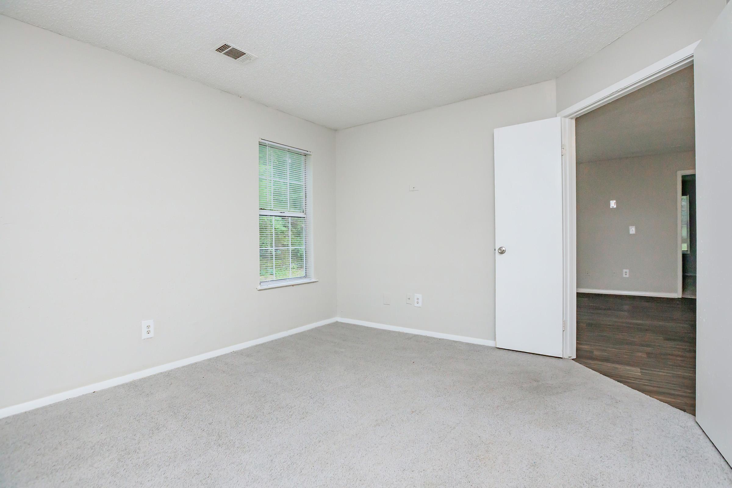 A vacant room with light gray walls and carpeted flooring. It features a window with blinds, allowing natural light to enter. One wall has an open doorway leading to another space. The room is uncluttered, creating a spacious and airy feel.