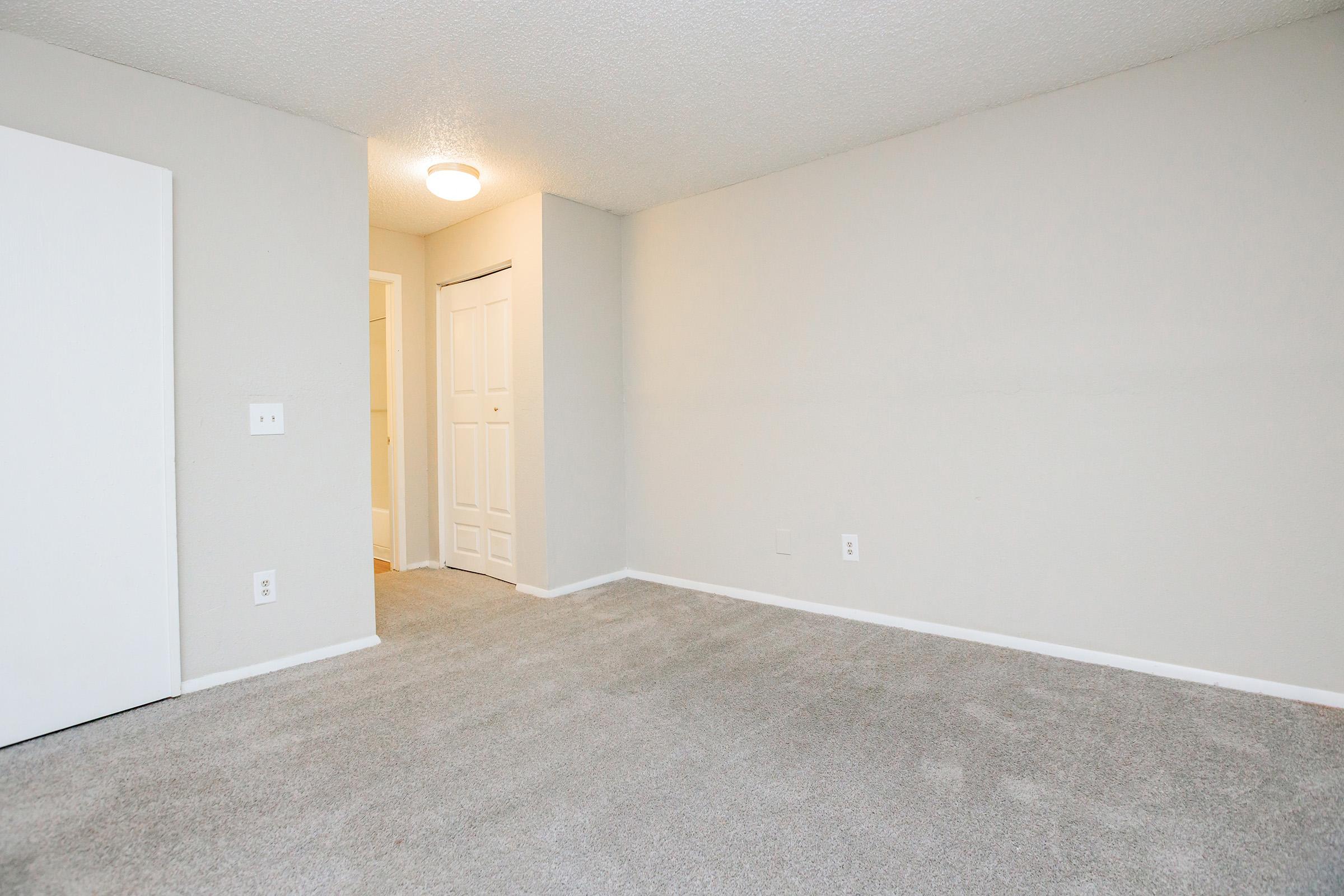 A spacious room with light gray carpet, painted beige walls, and a ceiling light. One wall features a white door that leads to a closet, while another wall shows an opening to a hallway. The room is empty, creating a bright and airy atmosphere, ideal for various uses or furnishings.