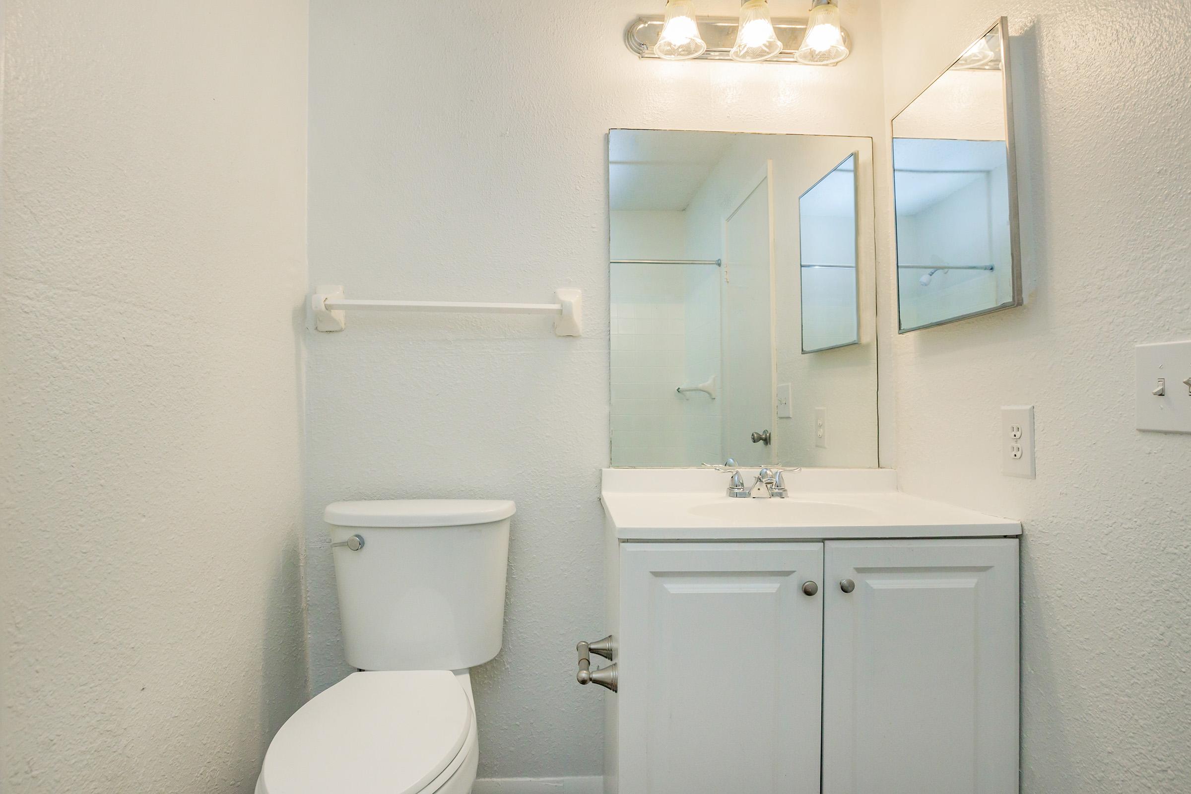 A clean, modern bathroom featuring a white toilet, a double sink vanity with a mirror above, and a shower area in the background. The walls are painted a light color, and there is a simple lighting fixture above the mirrors, creating a bright and airy atmosphere.