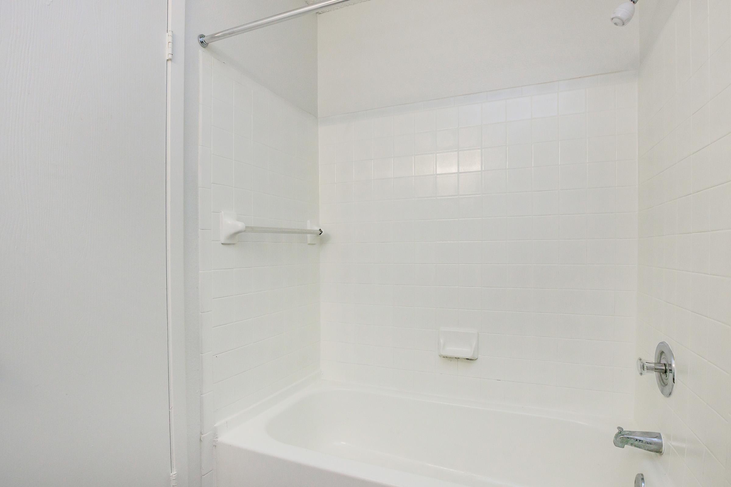 A clean, white-tiled bathroom with a bathtub, single towel rack, and minimal fixtures. The walls and floor are also white, creating a bright and spacious feel. Natural light enhances the simplicity of the design.