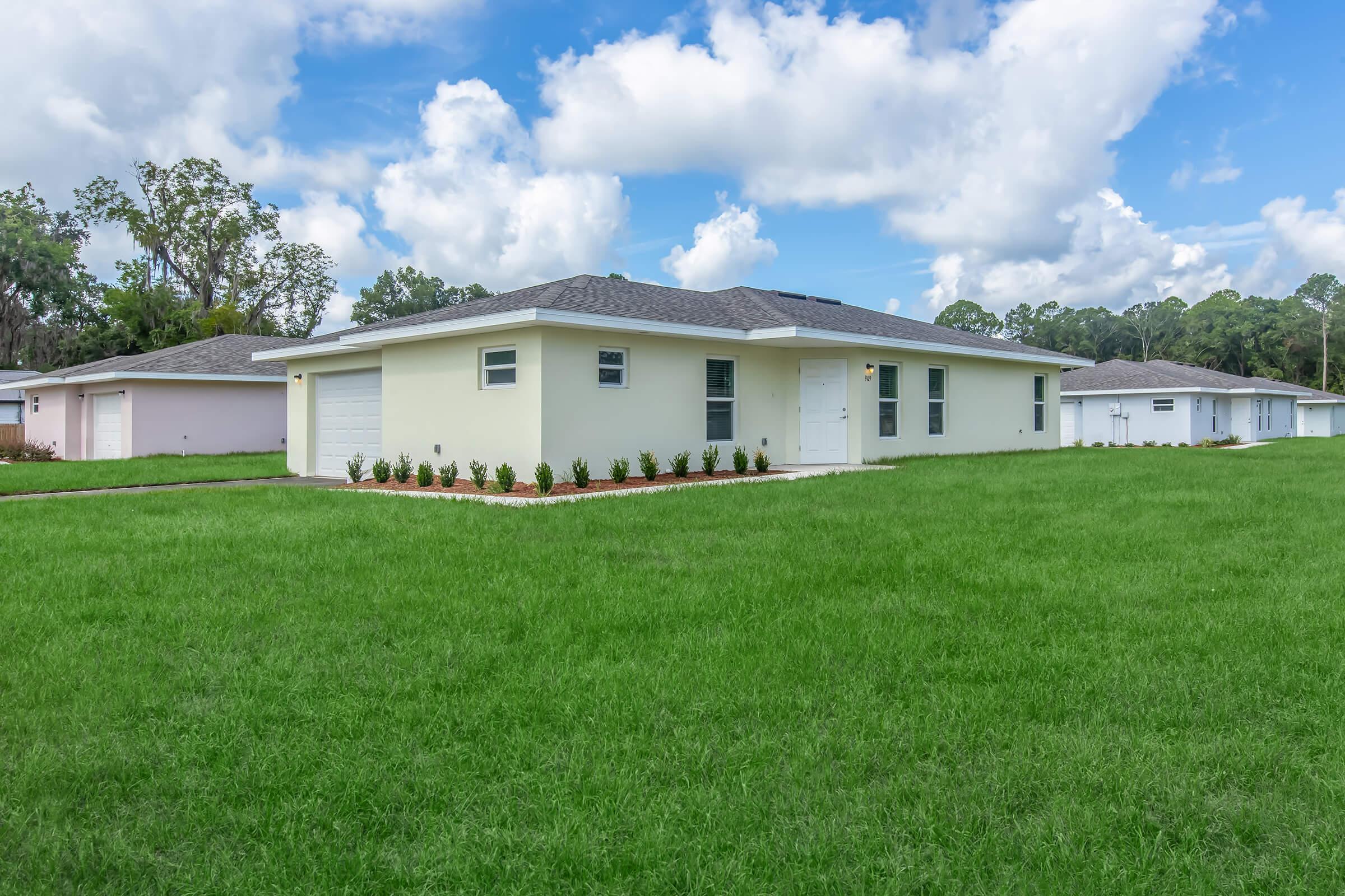 A single-story home with a light-colored exterior and a gray roof, set on a spacious green lawn. The front yard features a neatly landscaped area with small plants. There are partly cloudy blue skies in the background, enhancing the bright and inviting atmosphere of the scene.