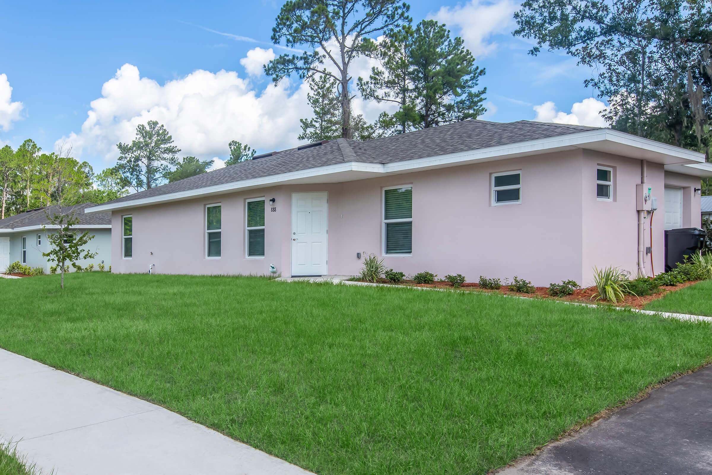 A single-story pink house with a well-manicured front lawn and a concrete walkway. The house features large windows, a white front door, and is surrounded by trees under a blue sky with scattered clouds.