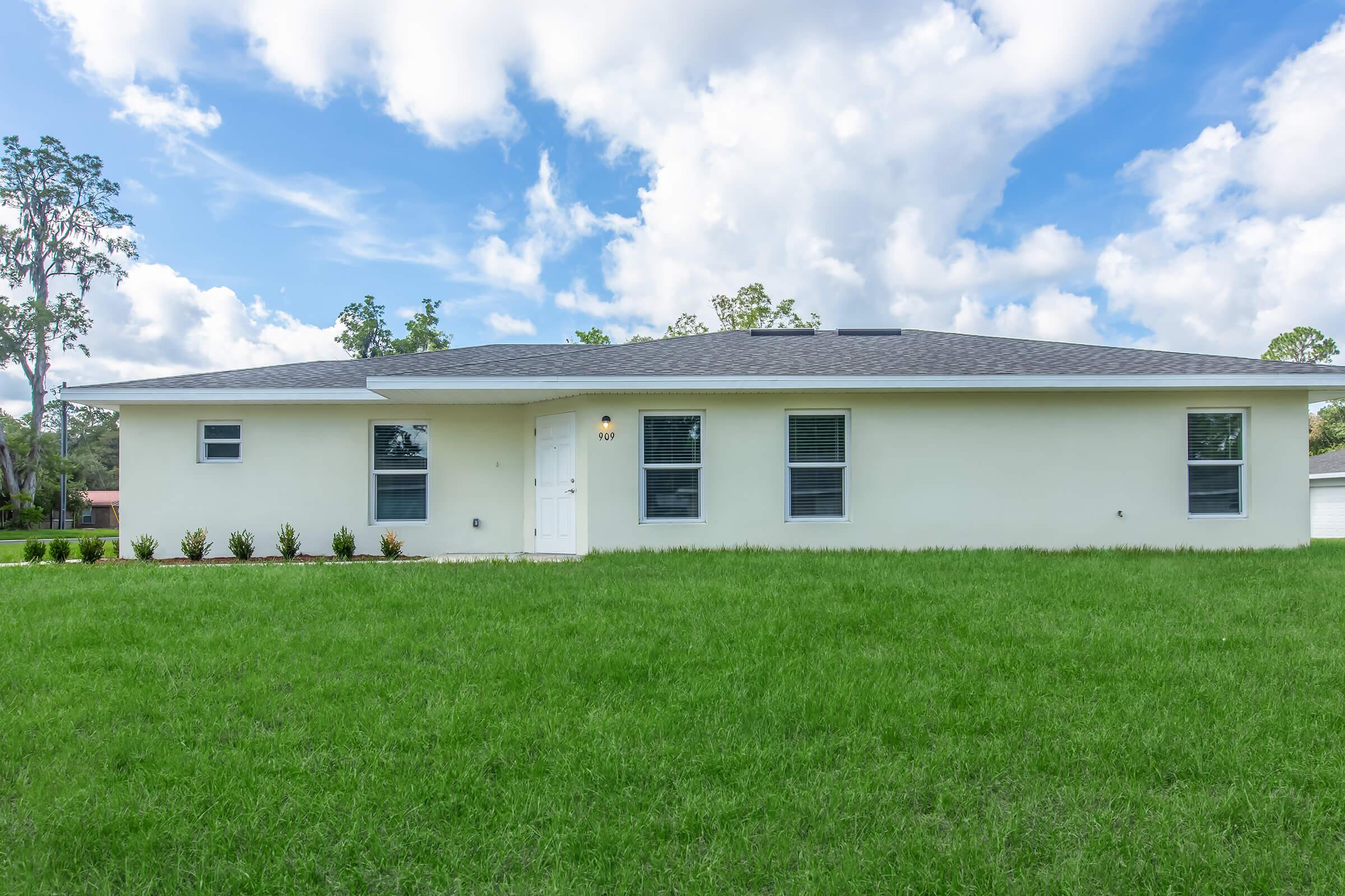 Single-story white house with a gray roof, featuring several windows on the front. The house is surrounded by a lush green lawn and is set against a backdrop of blue sky with fluffy white clouds.