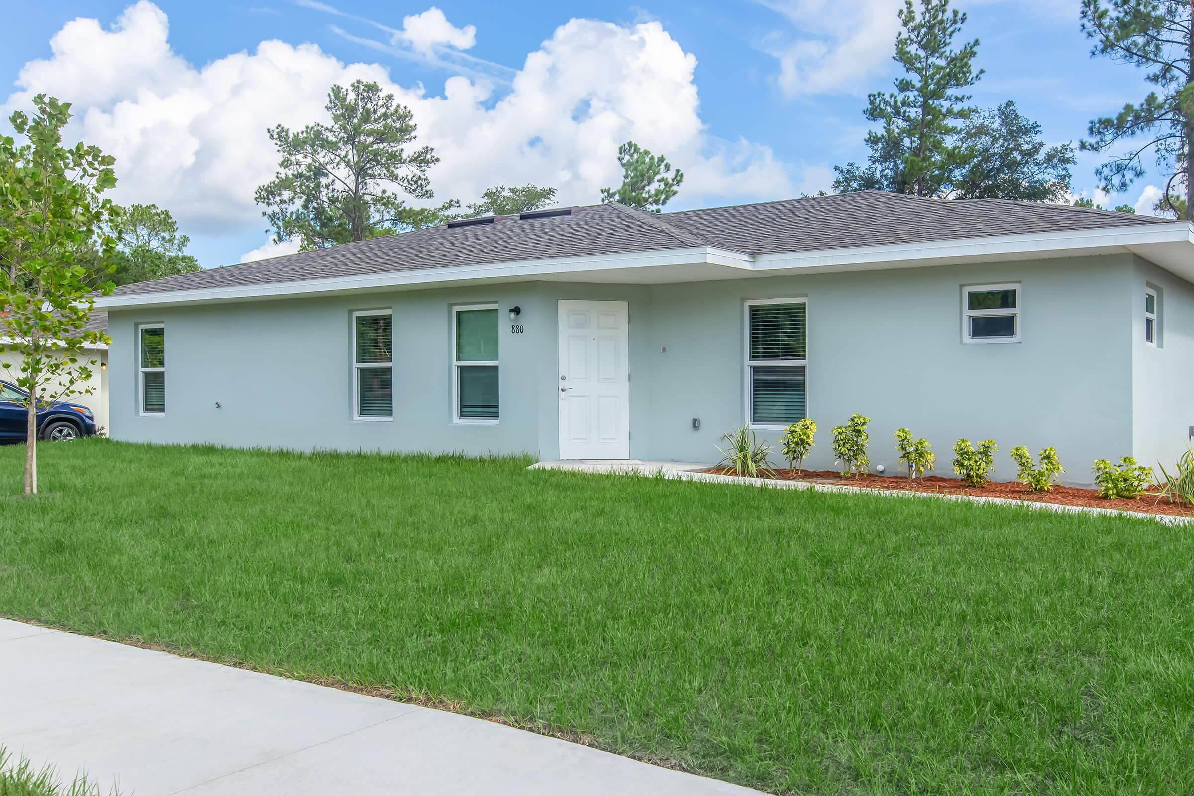 A modern single-story house with a light gray exterior, white front door, and large windows, surrounded by well-manicured grass and small shrubs. The house features a simple design with a flat roof, and there are trees and blue skies in the background. A concrete pathway leads to the entrance.