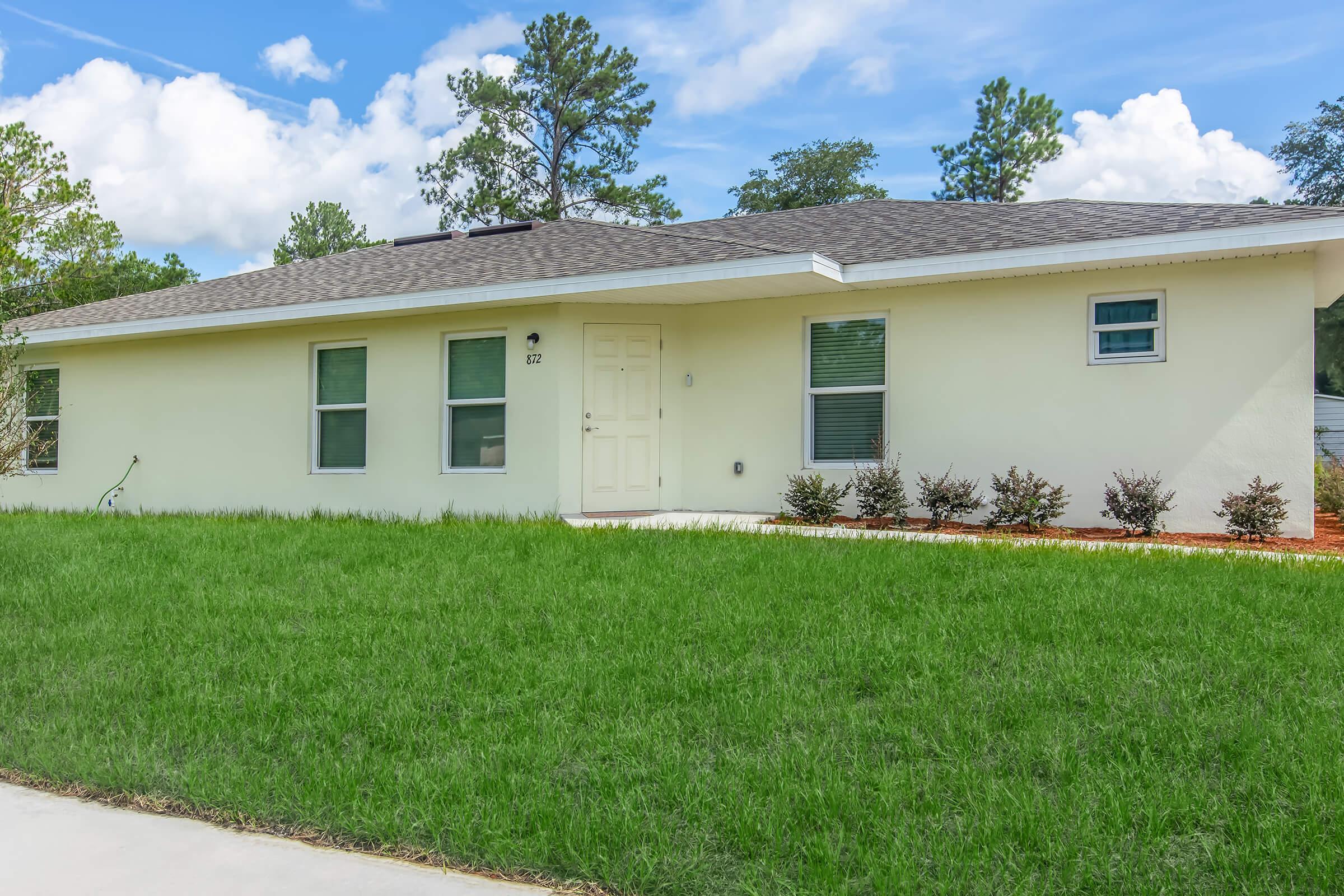 A single-story yellow house with a dark gray roof, featuring multiple windows and a white front door. The house is surrounded by green grass and small shrubs, with a clear blue sky and scattered clouds in the background. A concrete pathway leads up to the house.