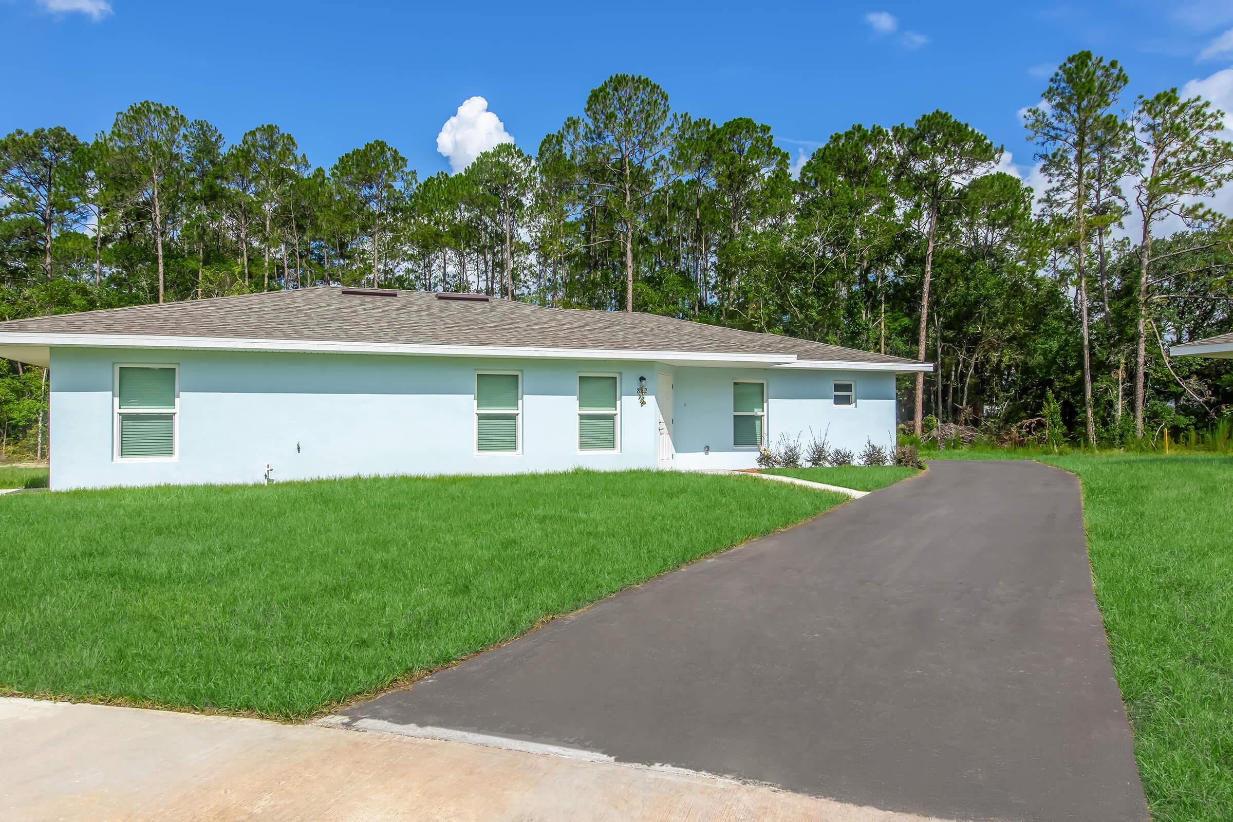 Single-story light blue house with a gray roof, situated on a green lawn. A driveway leads up to the house, surrounded by trees in the background under a clear blue sky.