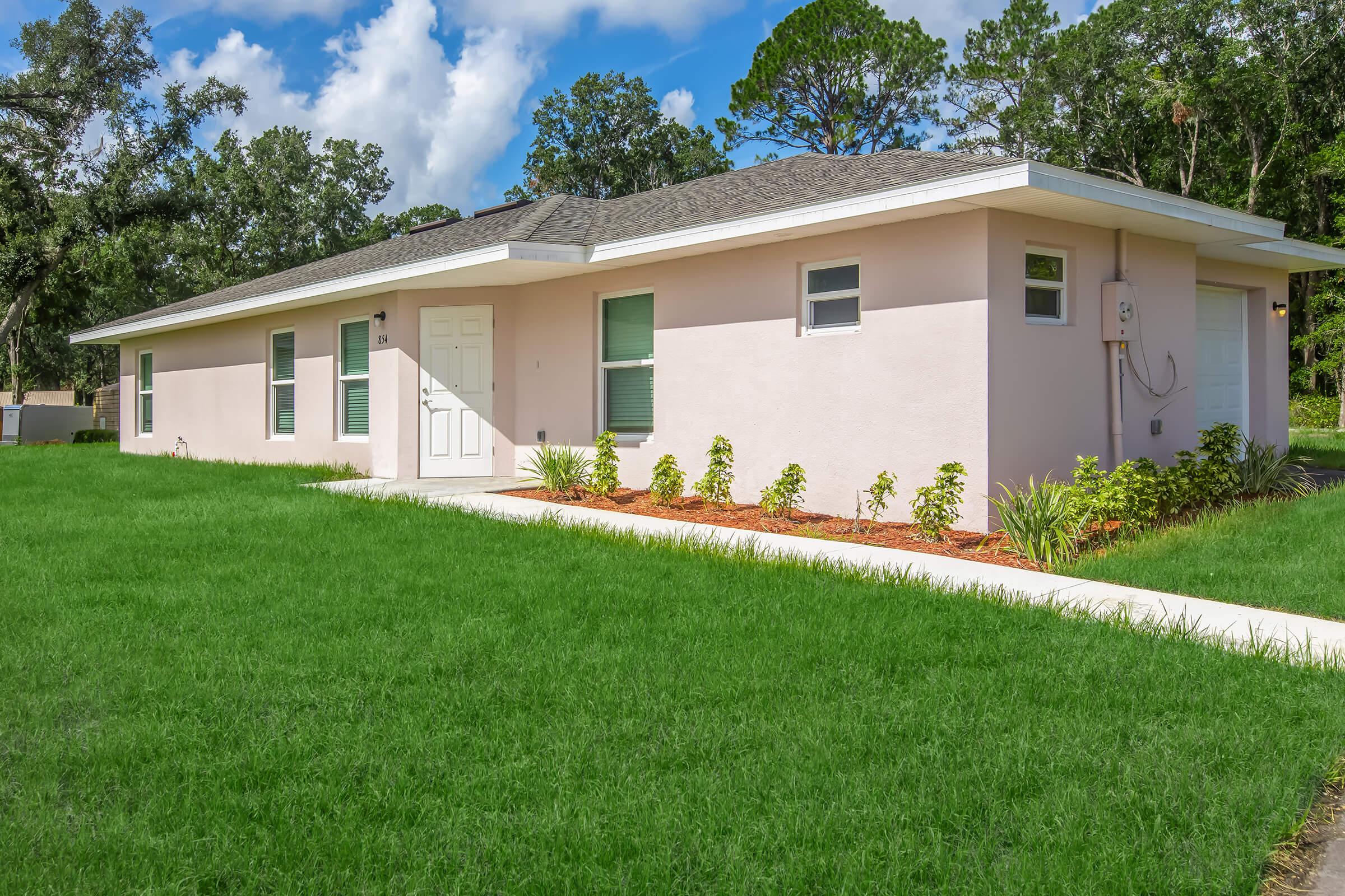 A modern single-story pink house with a sloped roof, featuring a front door and windows, surrounded by lush green grass and neatly maintained landscaping. The house has a driveway leading to a garage on the side, under a bright blue sky with scattered clouds.