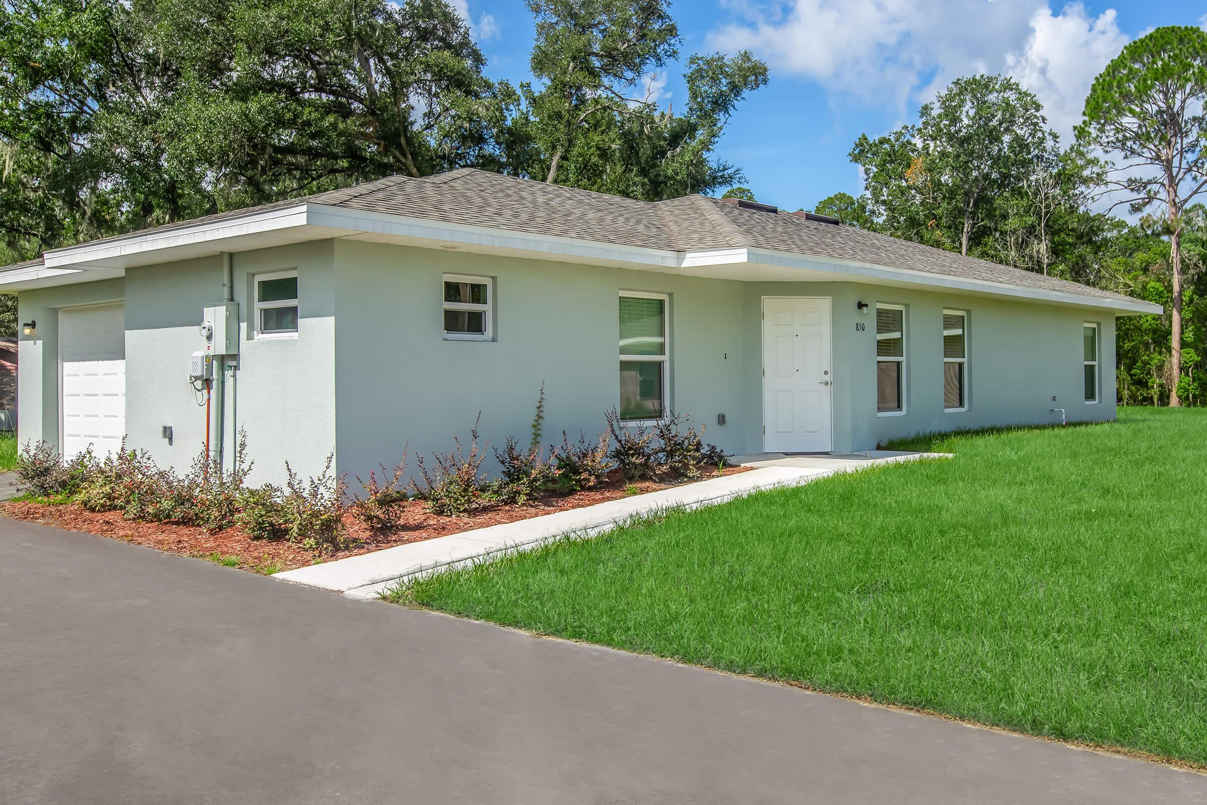 A single-story house with light blue exterior and a gray roof, surrounded by lush green grass. The front yard features a neatly trimmed lawn and flower beds. A paved driveway leads to the house, which has multiple windows and a front door. Trees and blue sky are visible in the background.