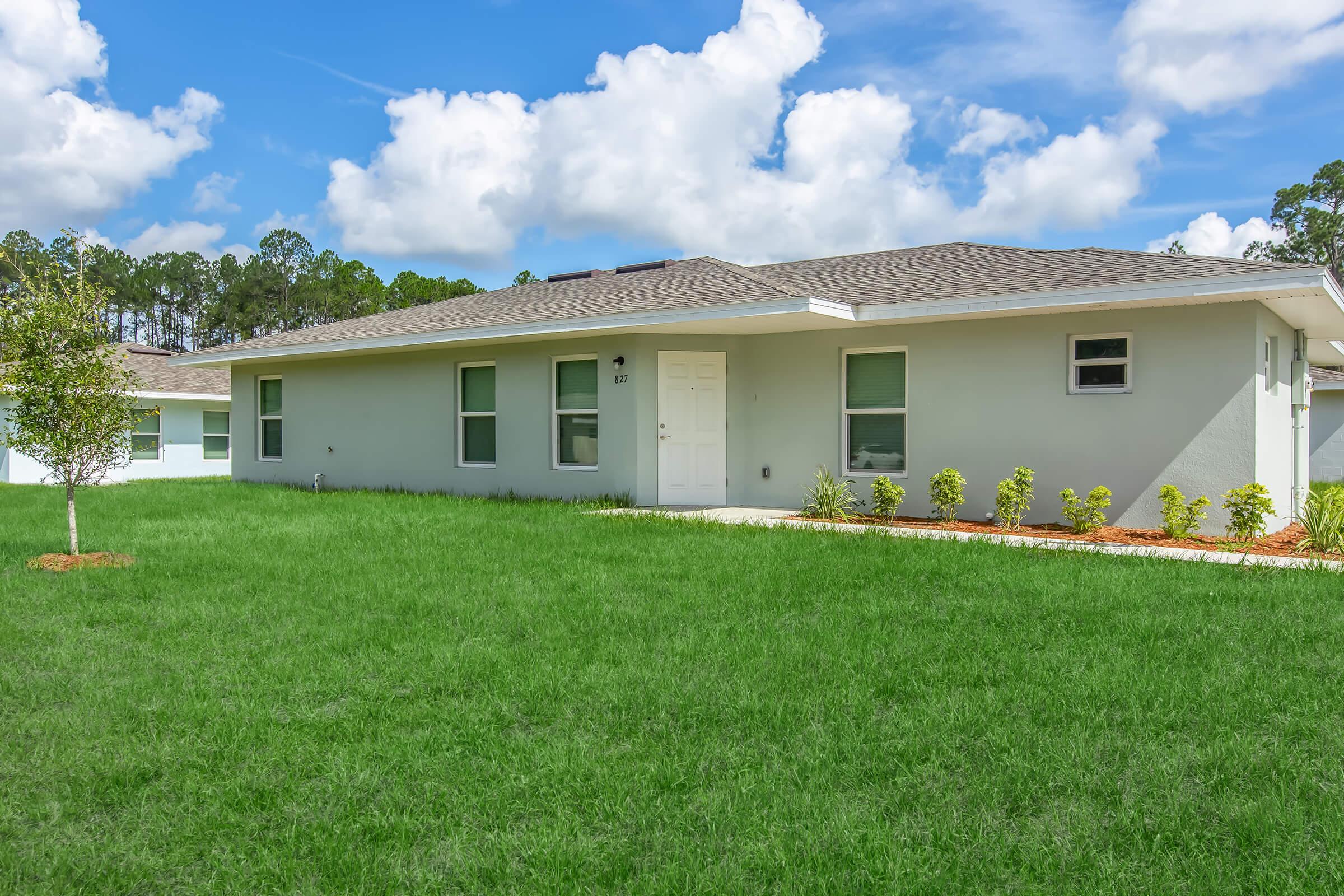 A modern single-story house with a light-colored exterior and a well-maintained front yard. The lawn is lush and green, featuring small shrubs along a path leading to the front door. Bright blue skies with fluffy white clouds create a sunny atmosphere.