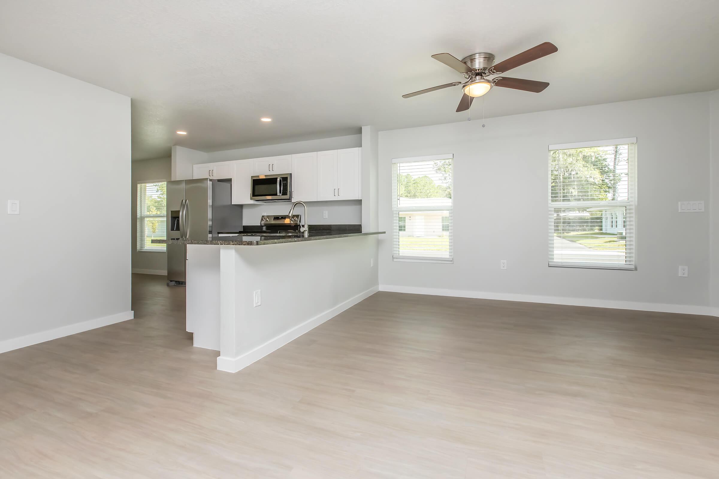 Spacious and modern open-concept living area with light wood flooring, featuring a ceiling fan and large windows. The adjacent kitchen showcases white cabinetry and stainless steel appliances. Natural light brightens the space, creating a welcoming atmosphere.