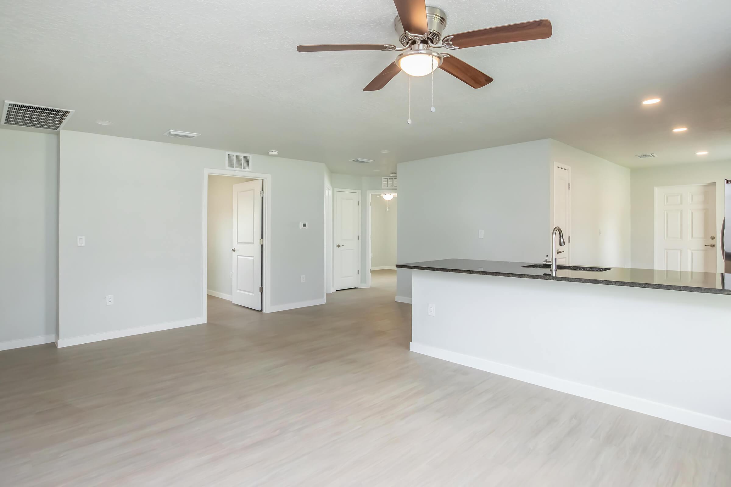 A spacious, modern living area featuring light-colored walls and wood-like flooring. There's a ceiling fan above, and an open-plan kitchen area on the left with a dark countertop. White doors lead to other spaces, creating a bright, inviting atmosphere.