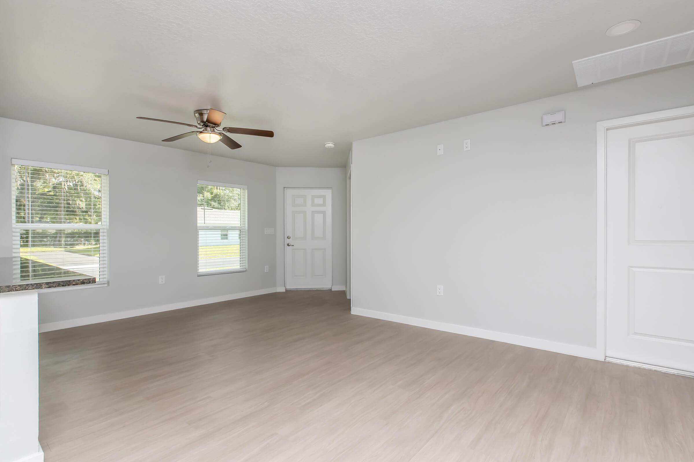 A spacious, empty living area featuring light-colored walls and a light wood floor. There is a ceiling fan in the center, a front door to the right, and two windows allowing natural light in. The overall ambiance is bright and clean, with a modern design.