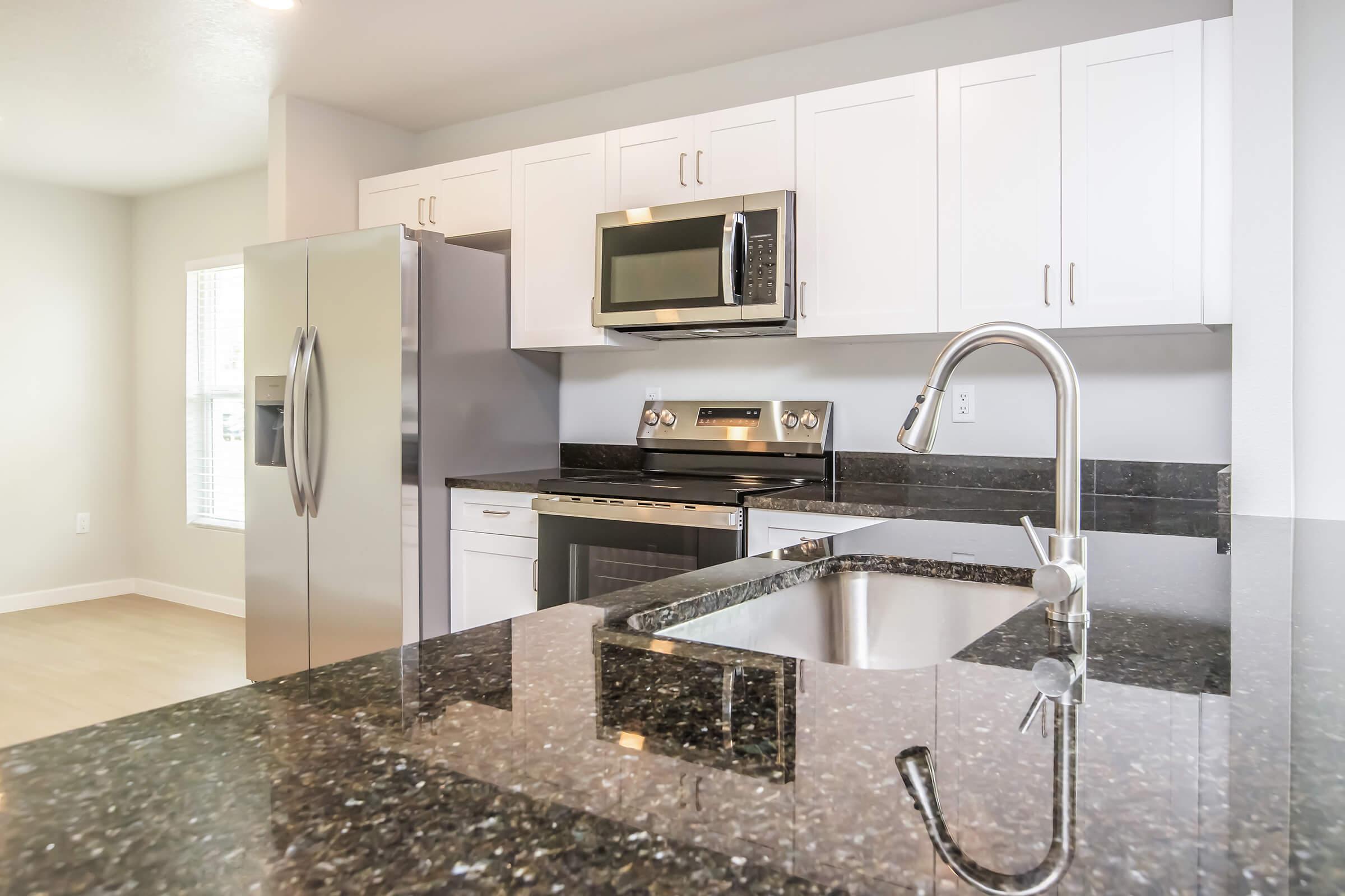Modern kitchen featuring stainless steel appliances, including a refrigerator and microwave, sleek dark countertops, and white cabinetry. The space is bright with natural light and has an open layout, complementing the contemporary design. A kitchen sink is visible on the island.