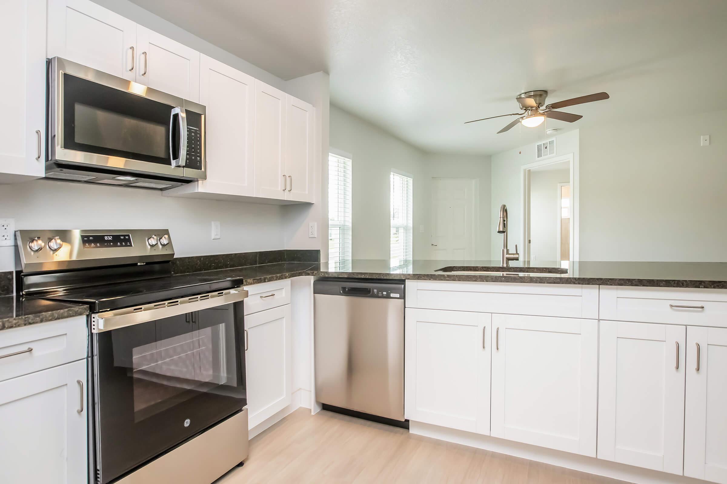 Modern kitchen featuring white cabinets, stainless steel appliances including a microwave and dishwasher, and a black countertop. The space has a ceiling fan, natural light from windows, and an open layout leading to another room. The flooring is light-colored, enhancing the bright atmosphere.