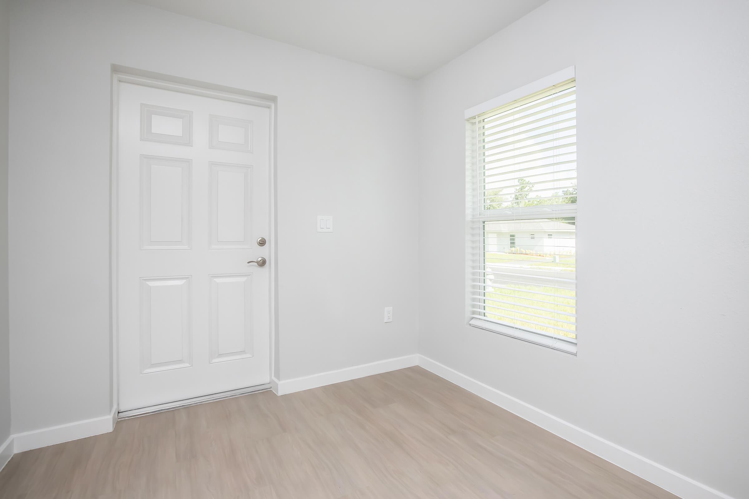 A light-filled room featuring a white door on one side and a window with blinds on the other. The walls are painted in a soft gray, and the floor is a light wood laminate. The space is empty and offers a welcoming, minimalistic atmosphere.
